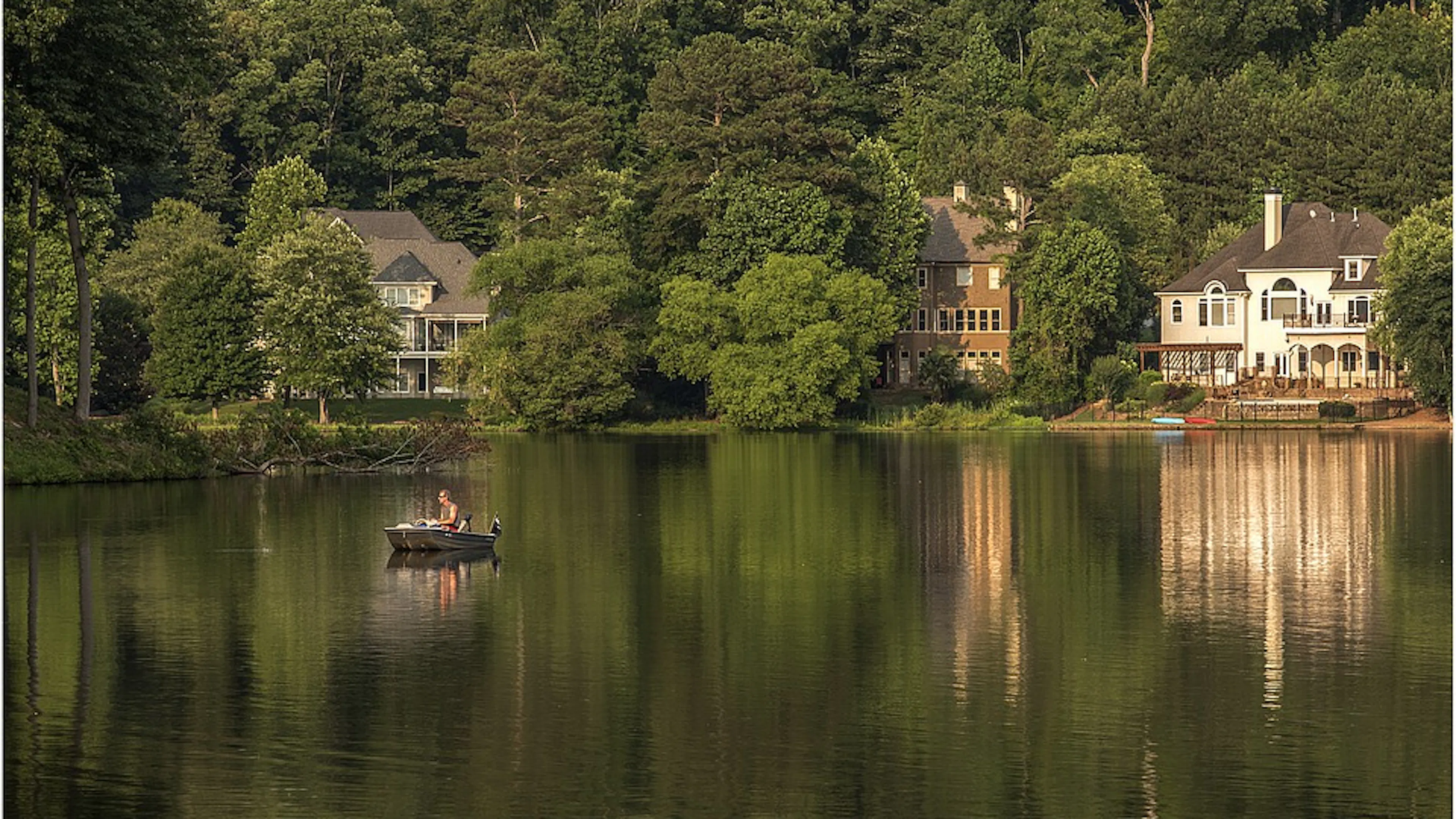 A lake surrounded by trees and houses with a lone boater on the water