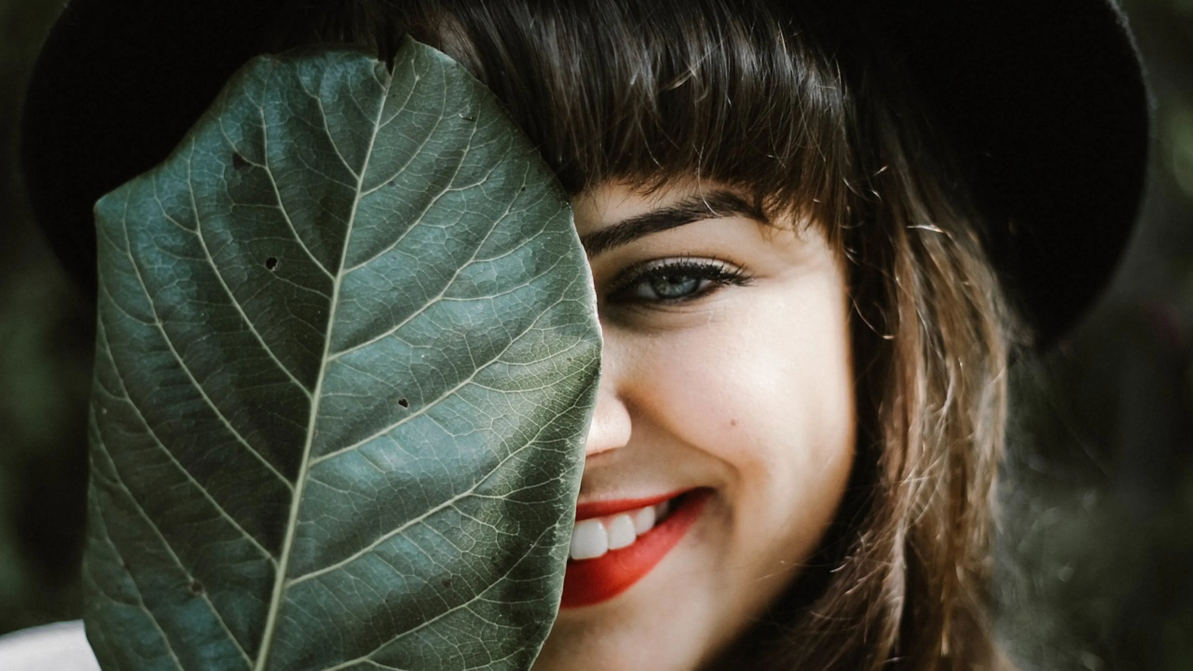 Woman holding leaf covering half her face as she smiles wearing a black hat