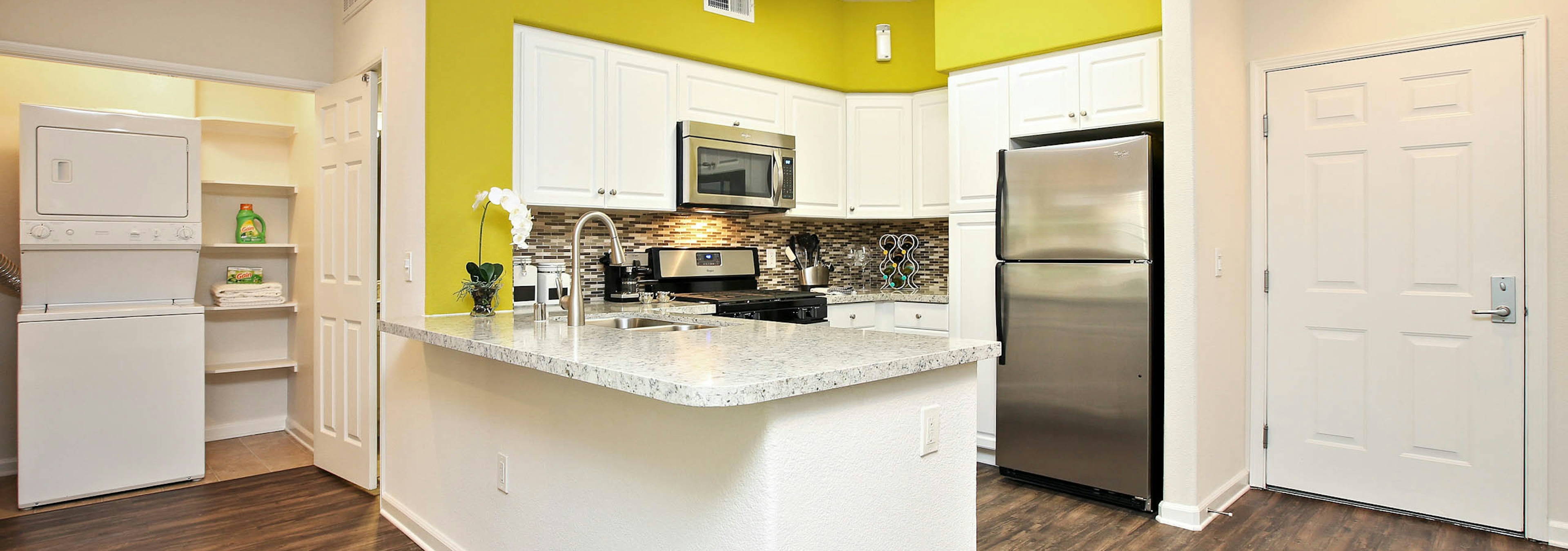 Interior of AMLI Warner Center apartment kitchen with white cabinetry, stainless steel appliances and peek into laundry area