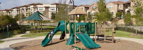 Playground with green slides on a bed of woodchips with a blue sky, trees and apartment building facade in the background