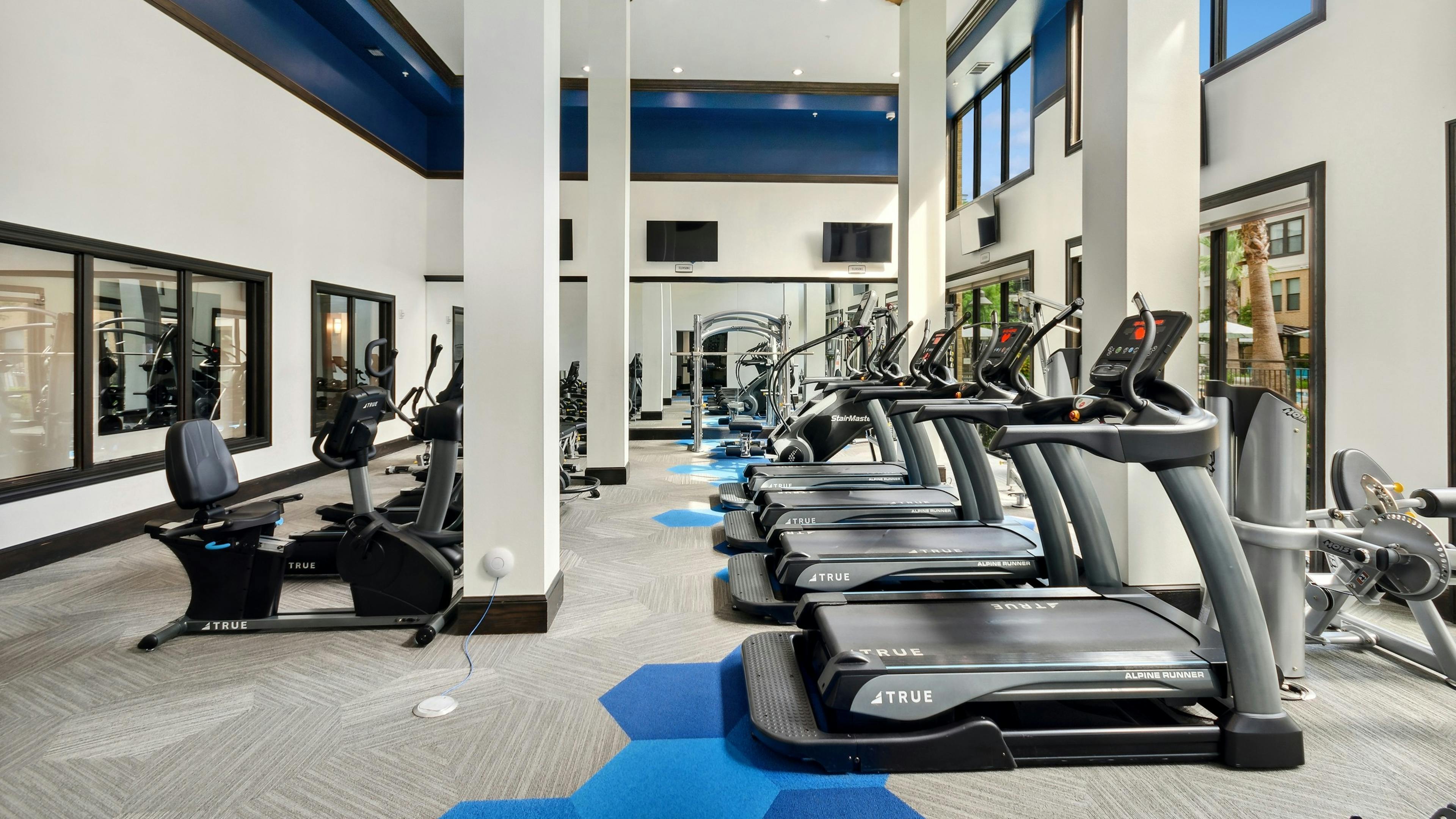 AMLI Campion Trail fitness center with treadmills and cardio machines and mounted TVs and large two-story windows