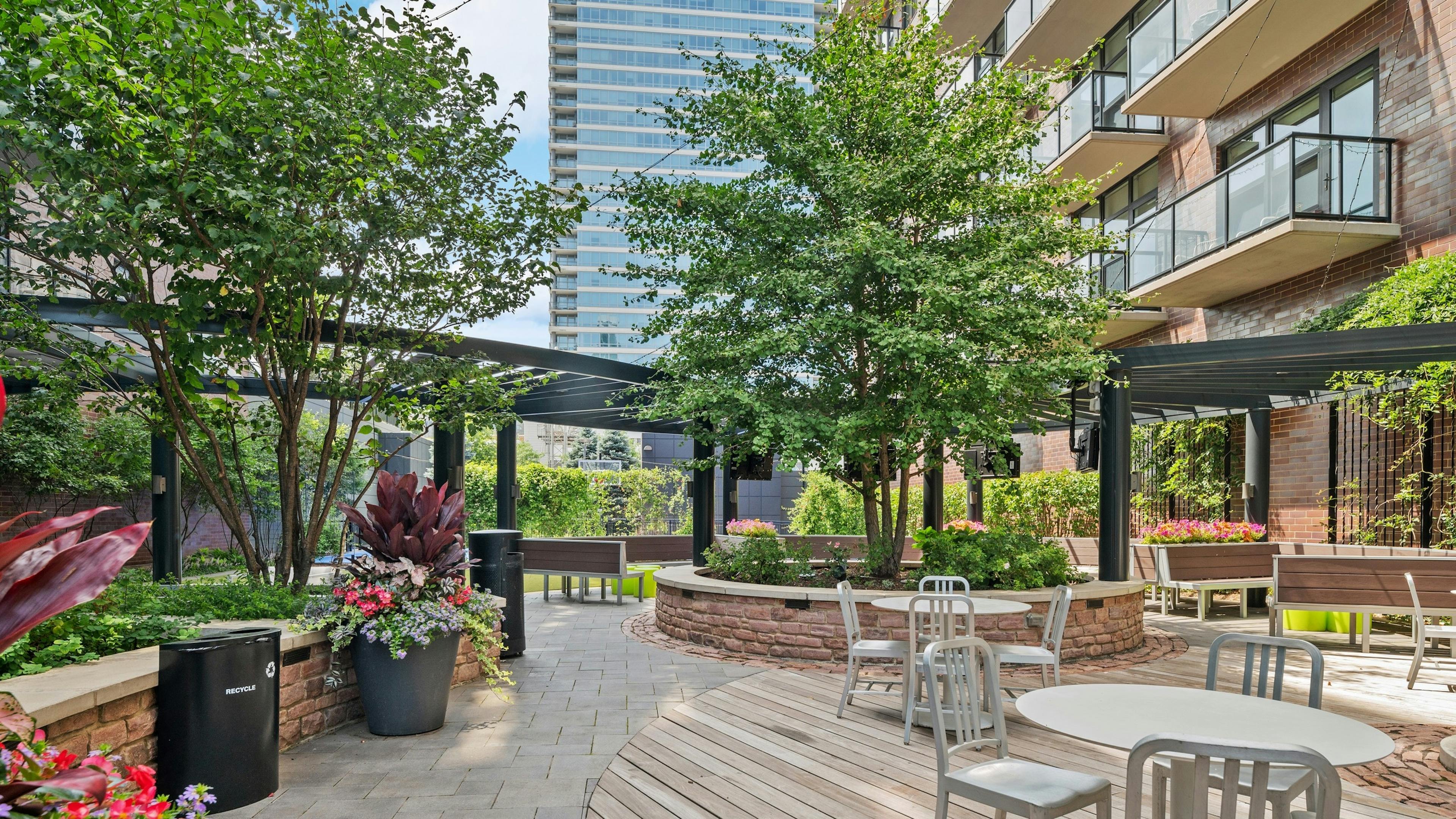 Daytime view of AMLI Lofts courtyard lounge with gray dining tables and wood bench seating surrounded by lush greenery