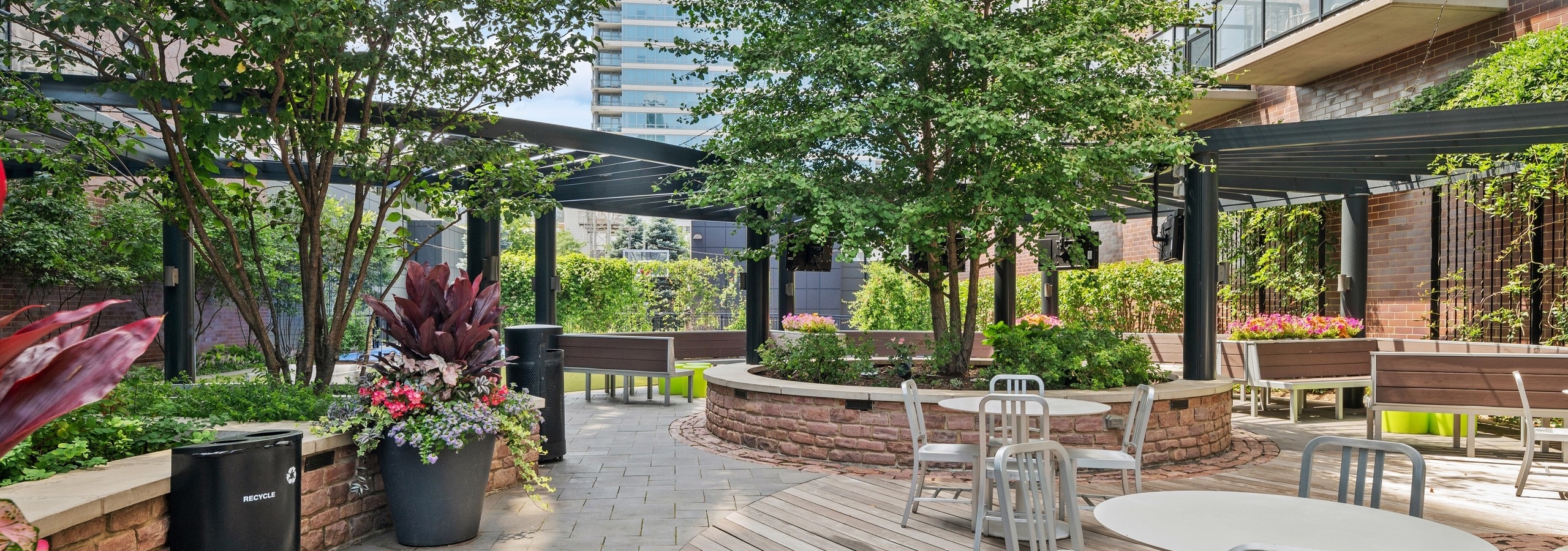 Daytime view of AMLI Lofts courtyard lounge with gray dining tables and wood bench seating surrounded by lush greenery