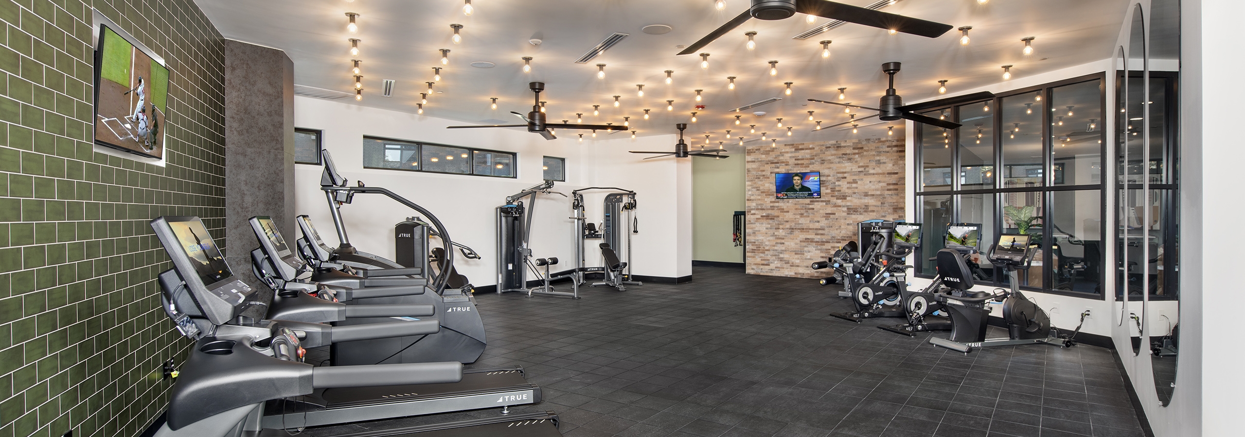 Fitness Center at AMLI Flatiron with gray floor and cardio equipment on green tile wall and ceiling fans and bulb lights above
