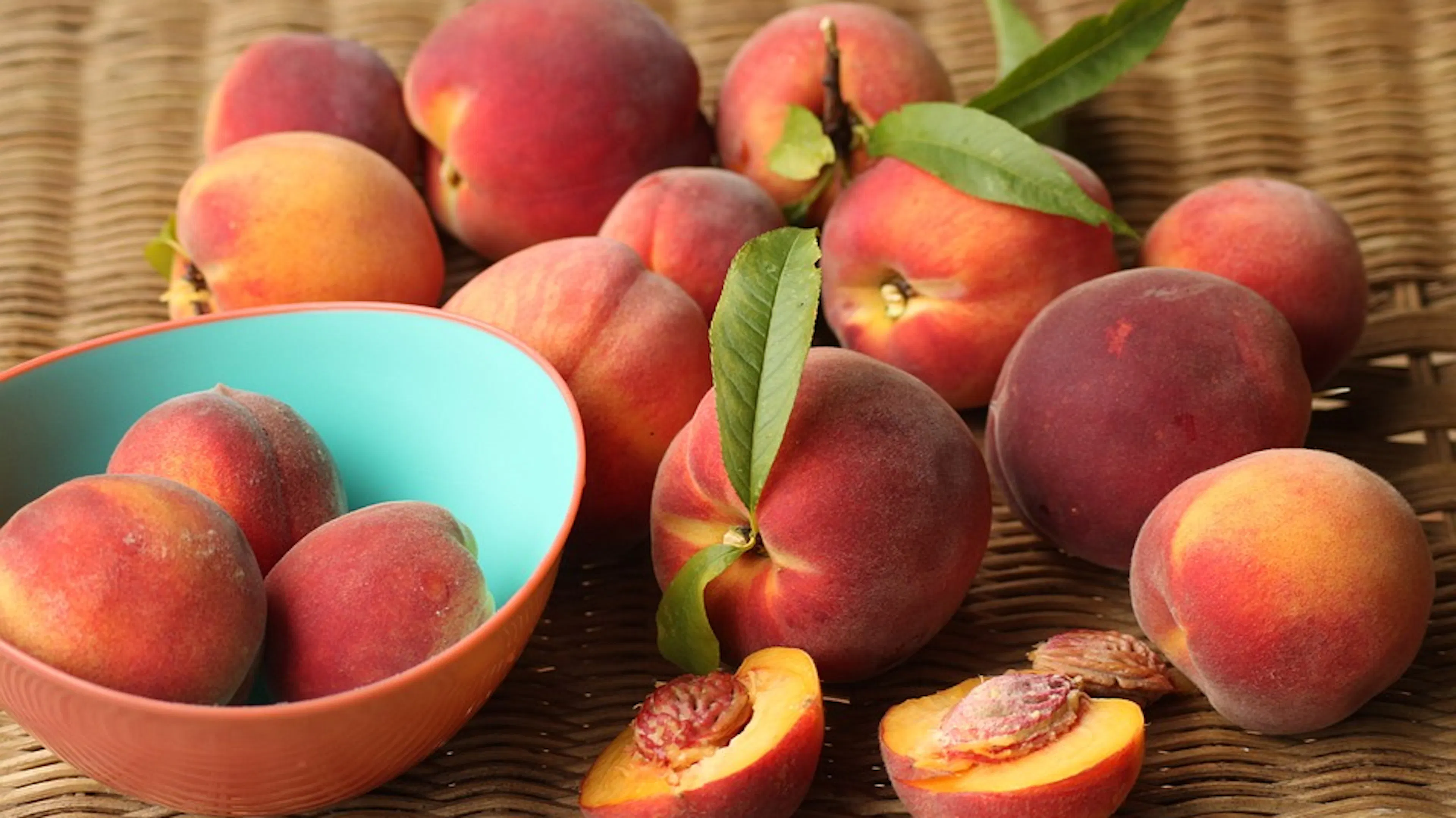 A bushel of peaches strewn out over a wicker basket and some in a bowl