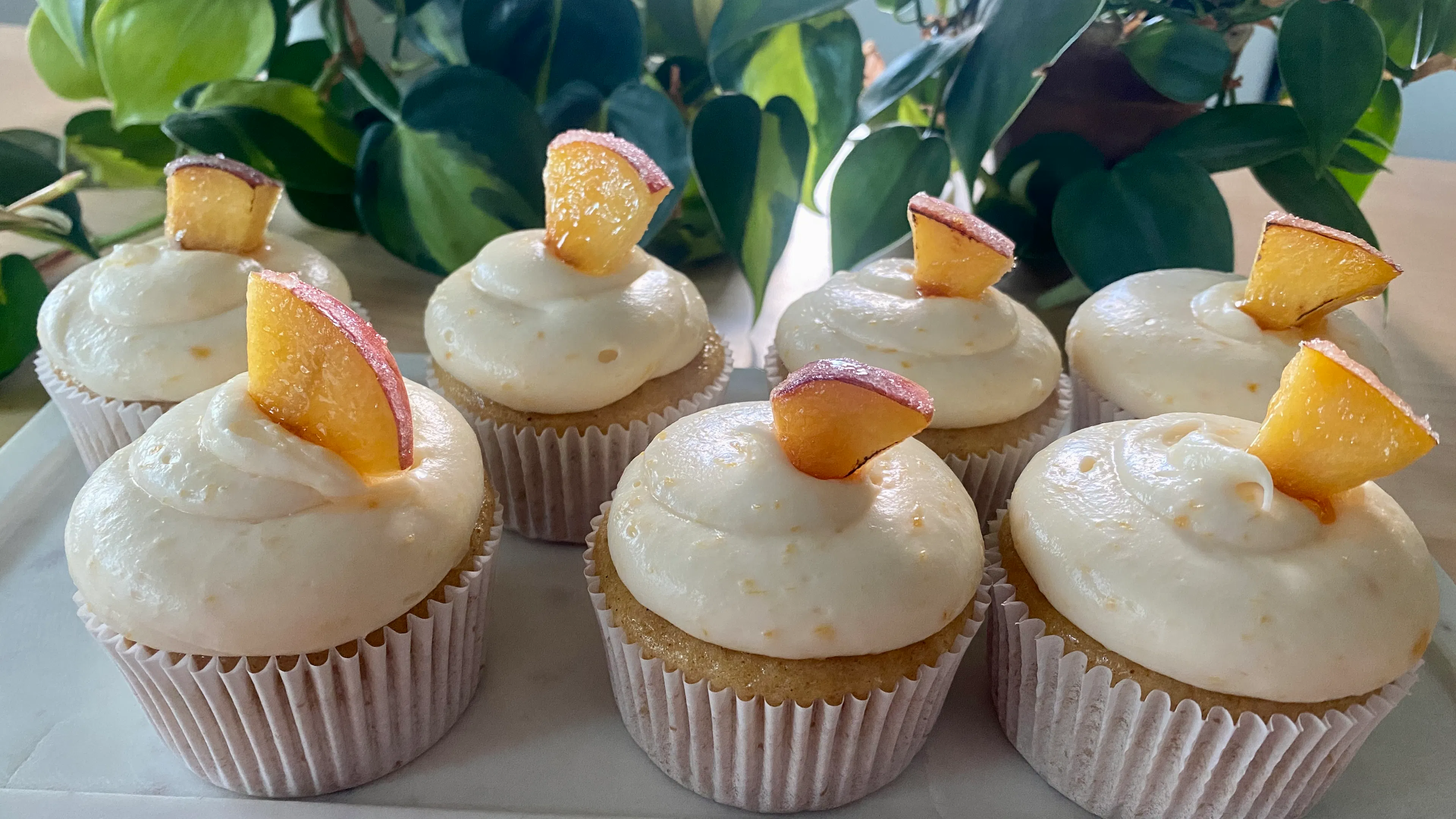Seven white cake cupcakes with cream cheese frosting and peach slices on top served on a white marble platter