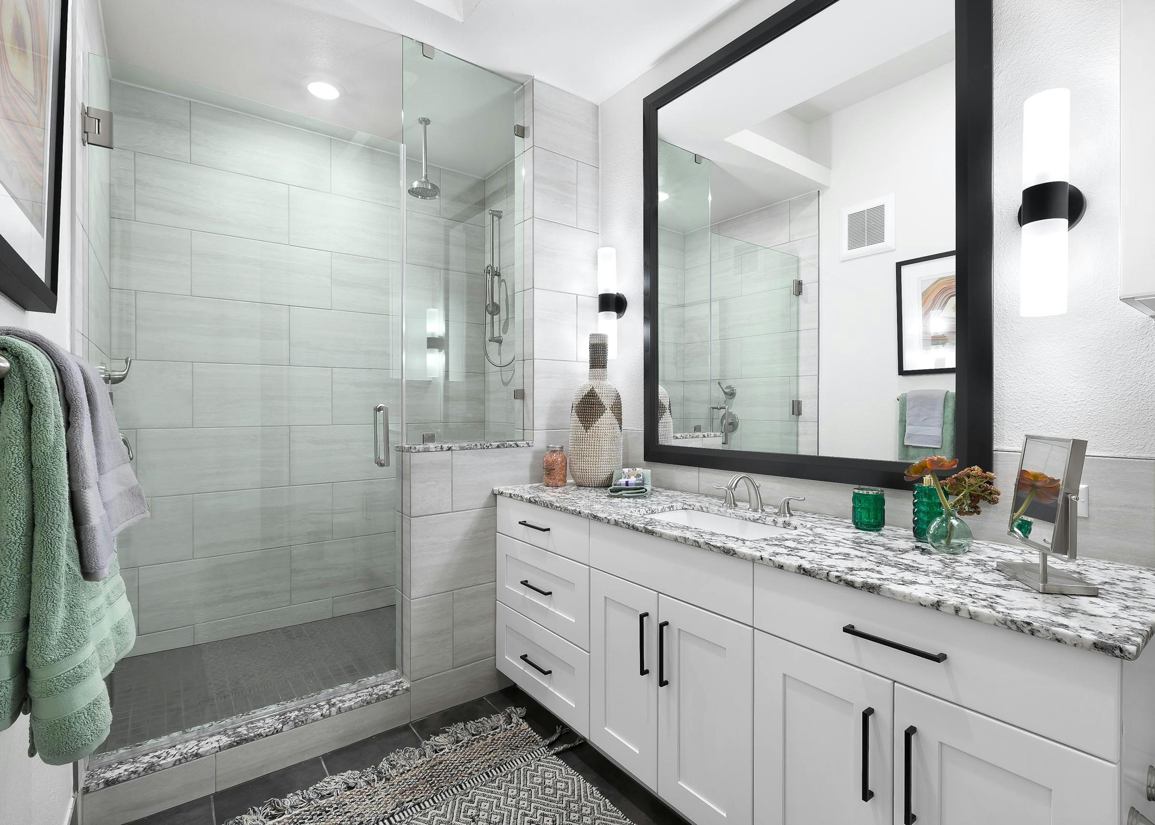 Interior view of AMLI Lakeline bathroom with white cabinetry with large mirror above and a tile and glass walk in shower with