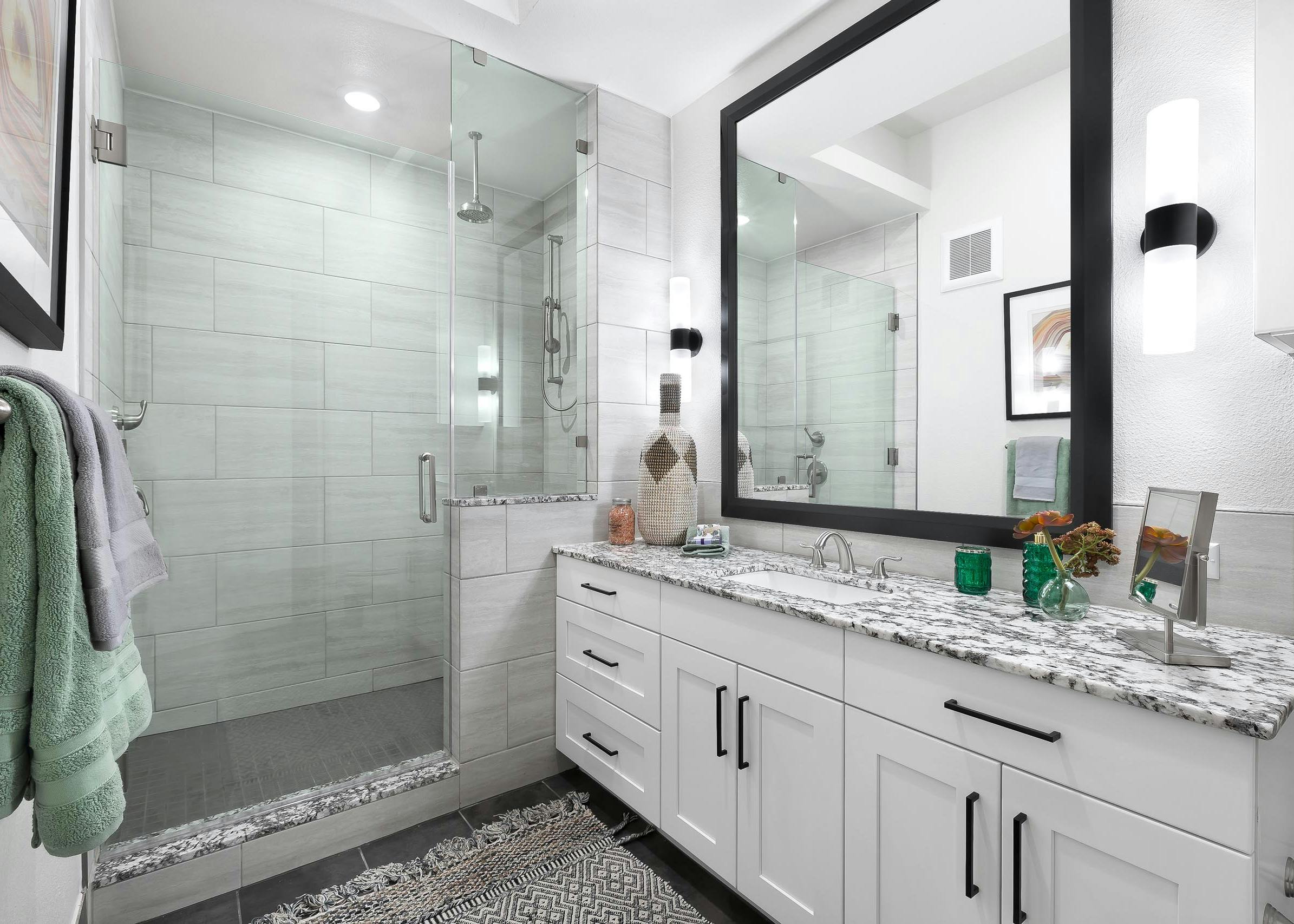 Interior view of AMLI Lakeline bathroom with white cabinetry with large mirror above and a tile and glass walk in shower with