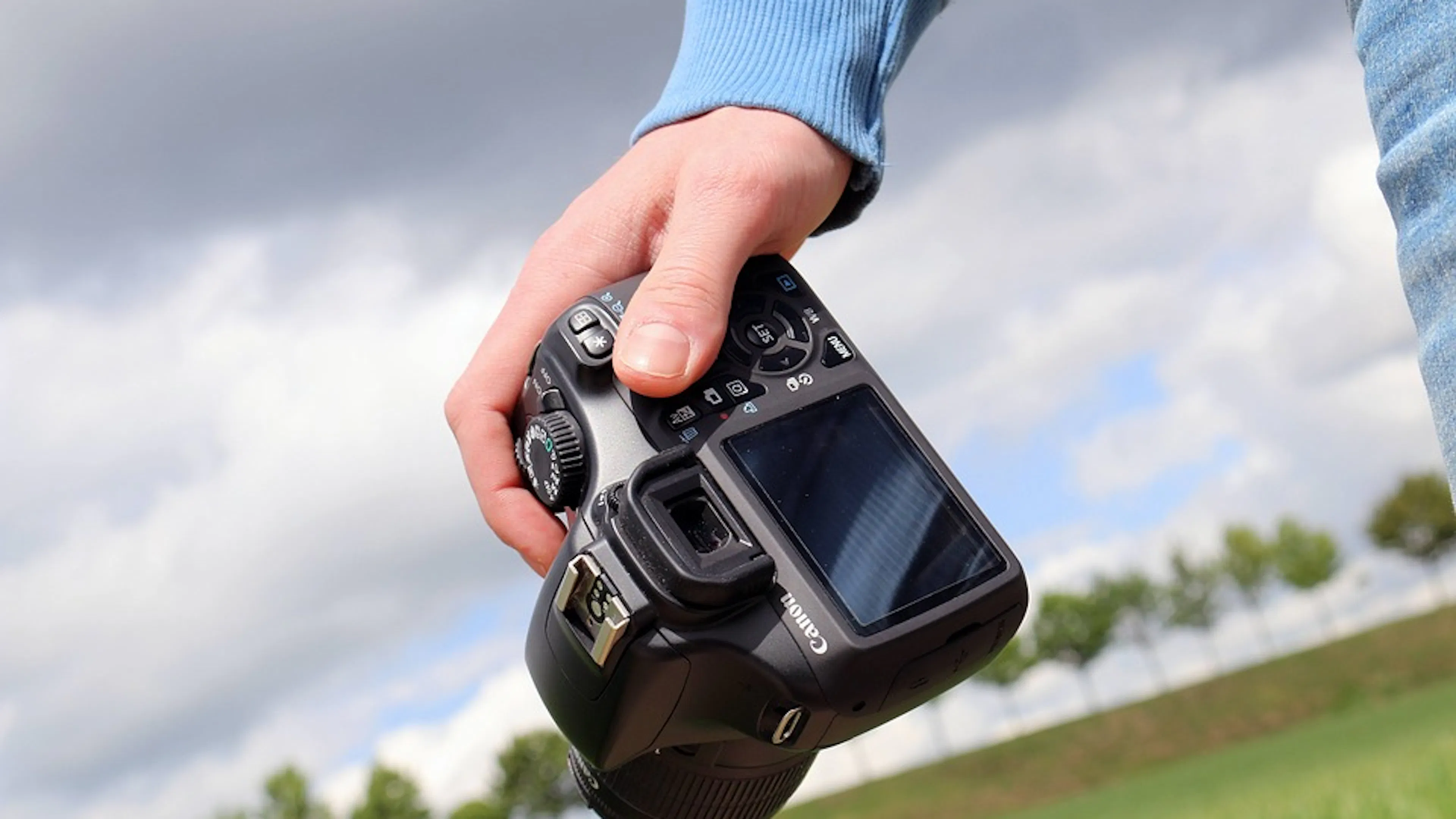 A hand holding a canon camera outdoors with cloudy sky and trees in background