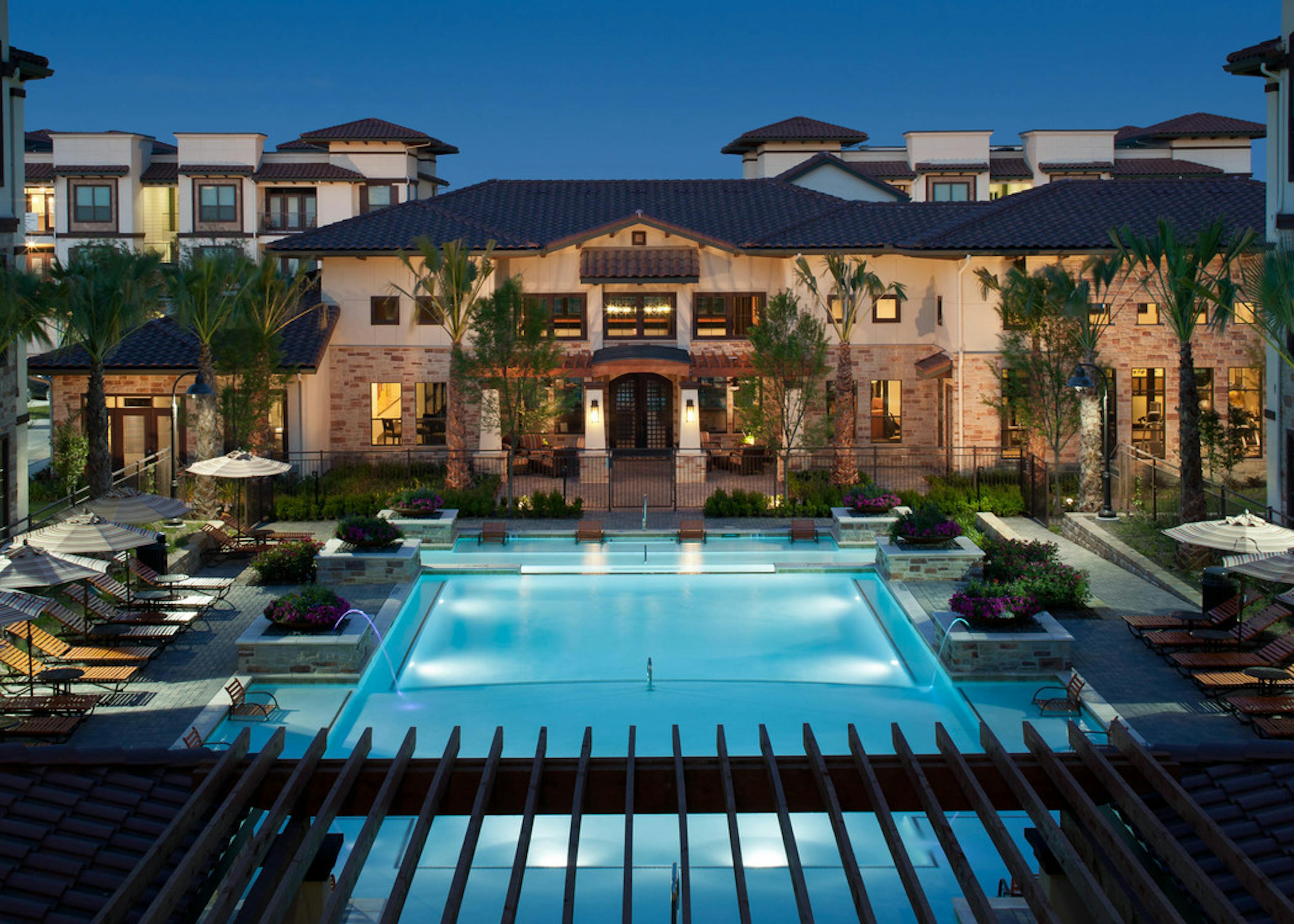 Nighttime view of AMLI at Escena apartments landscaped swimming pool area with lounge seating and umbrellas and palm trees