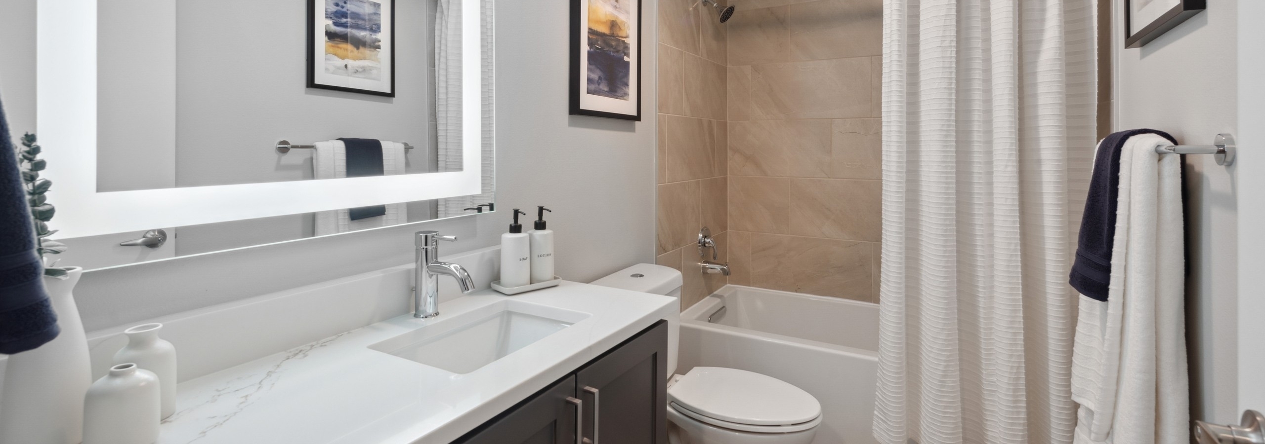 Bathroom at AMLI 535 with lighted mirror and white countertops with dark wood cabinets with earth tone tile tub surround