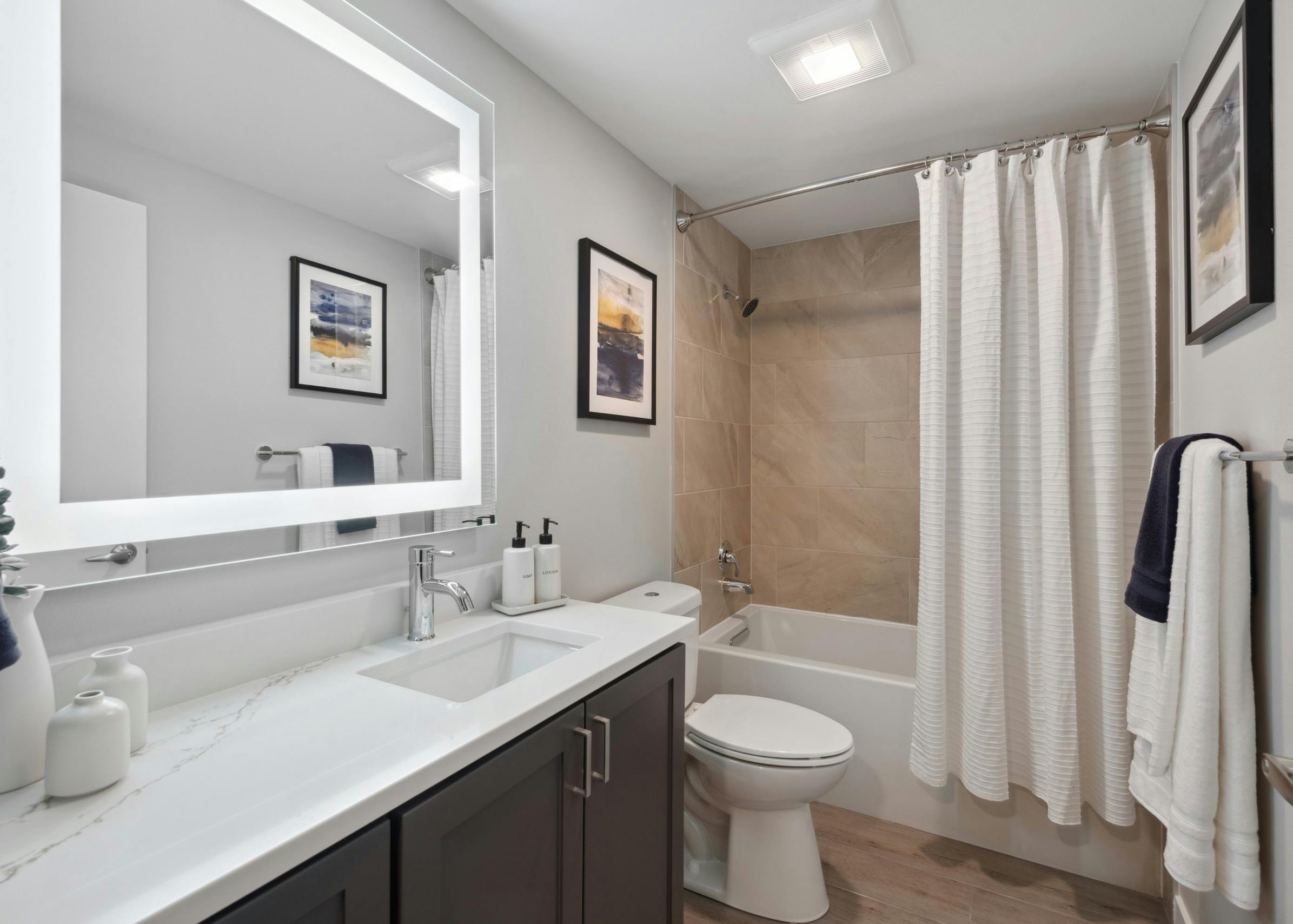 Bathroom at AMLI 535 with lighted mirror and white countertops with dark wood cabinets with earth tone tile tub surround