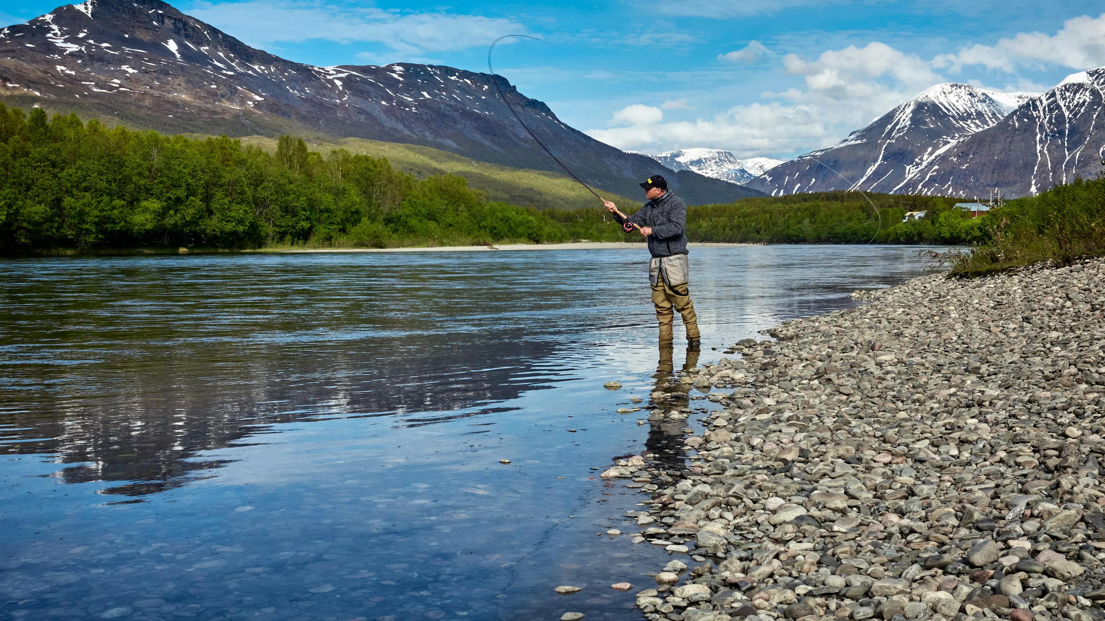A fly fisherman standing on the shore of an alpine lake surrounded by snow capped mountains