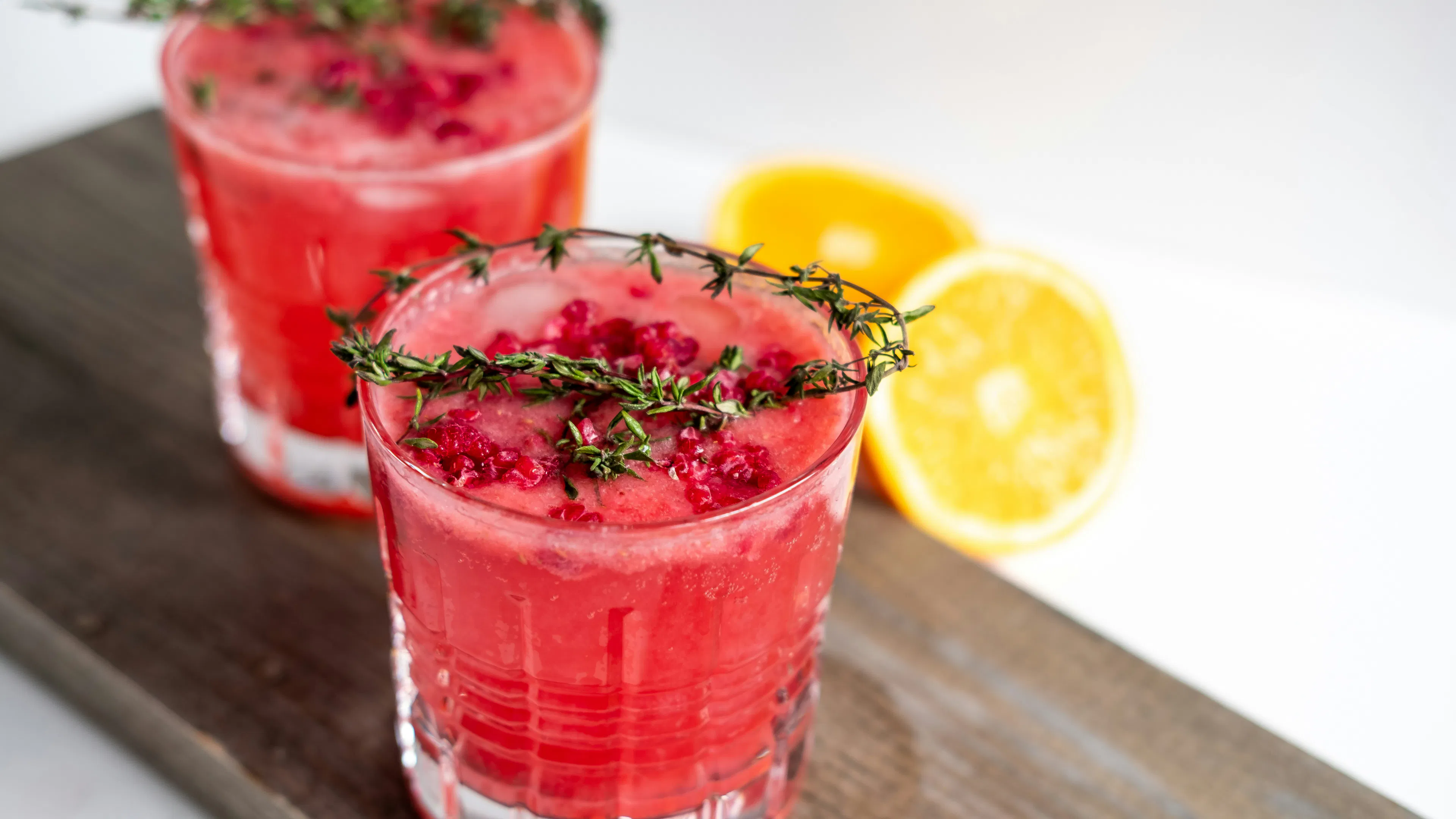 Two pink mocktails topped with rosemary on a wooden serving board beside some sliced lemons