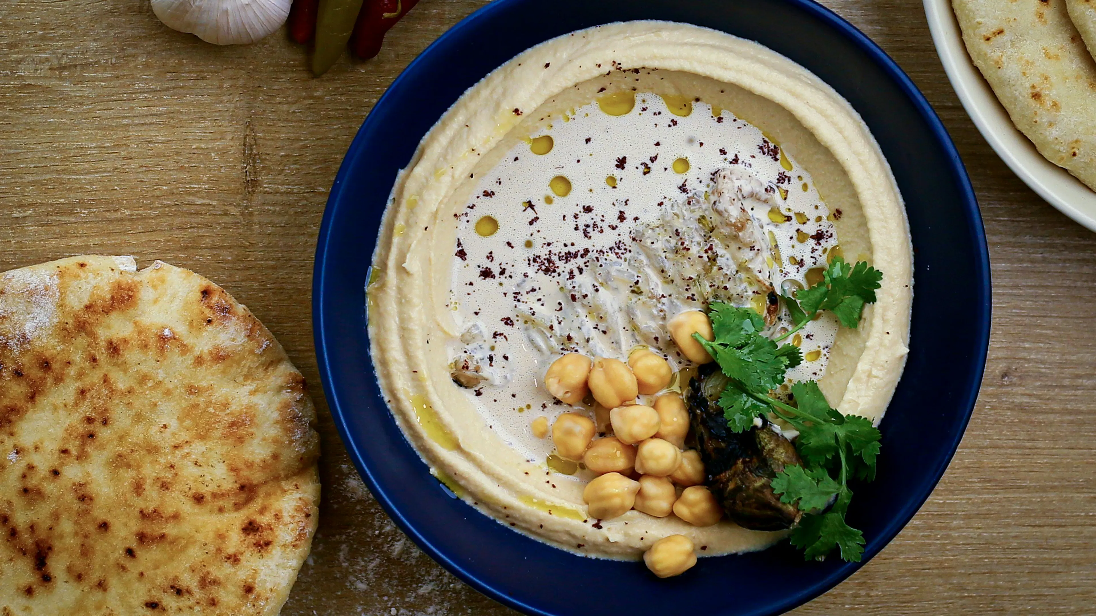 A plate of hummus garnished with chickpeas and parsley on a wooden table with some bread and garlic cloves sprinkled around for decoration