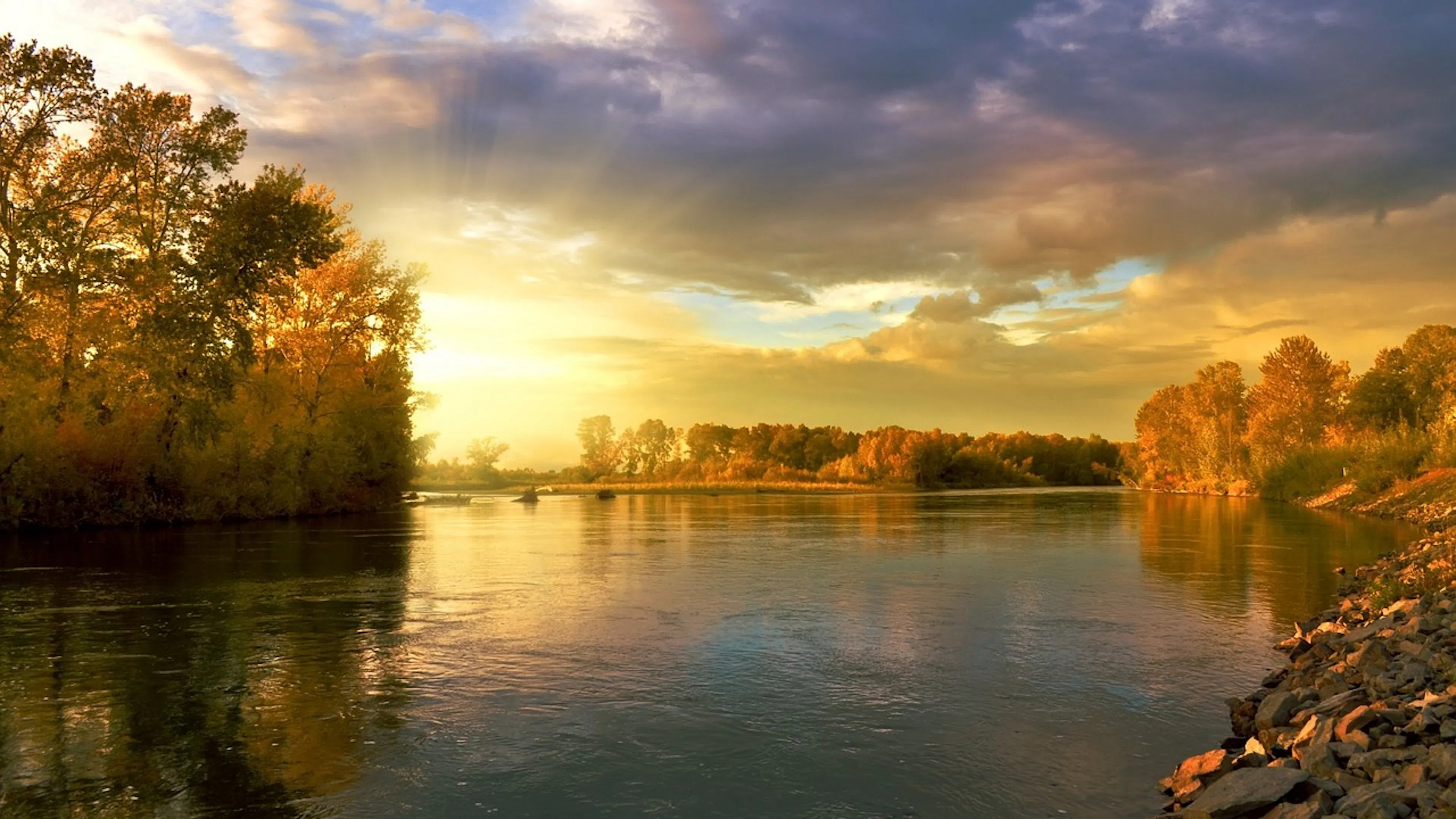 A wide calm river surrounded by trees with golden leaves as the sun sets behind them 