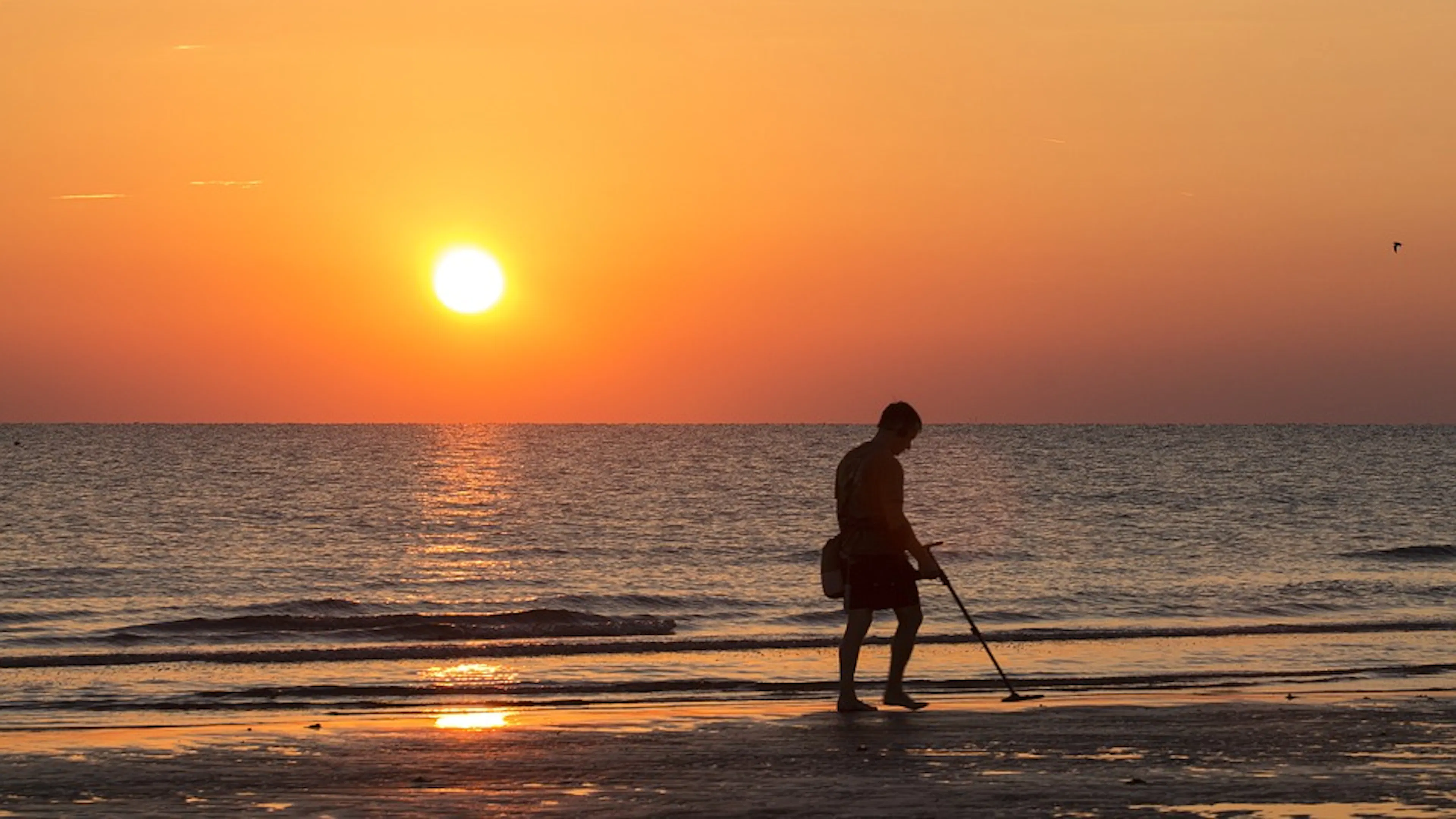 A man with a metal detector on the beach at sunrise with calm waters and clear skies in the background