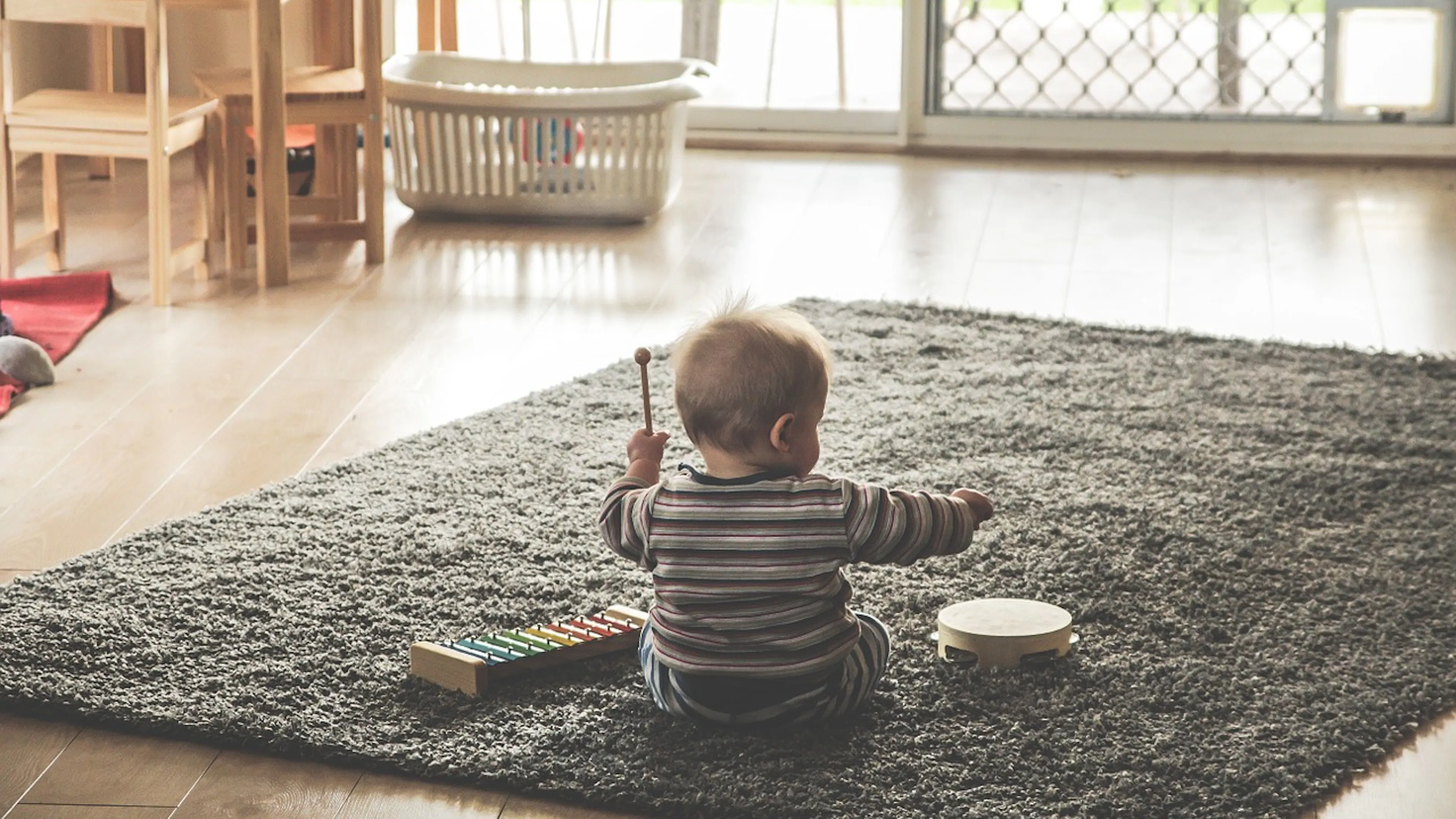 Back of baby in striped shirt holding mallet and playing with xylophone and tambourine on gray area rug on wood floor