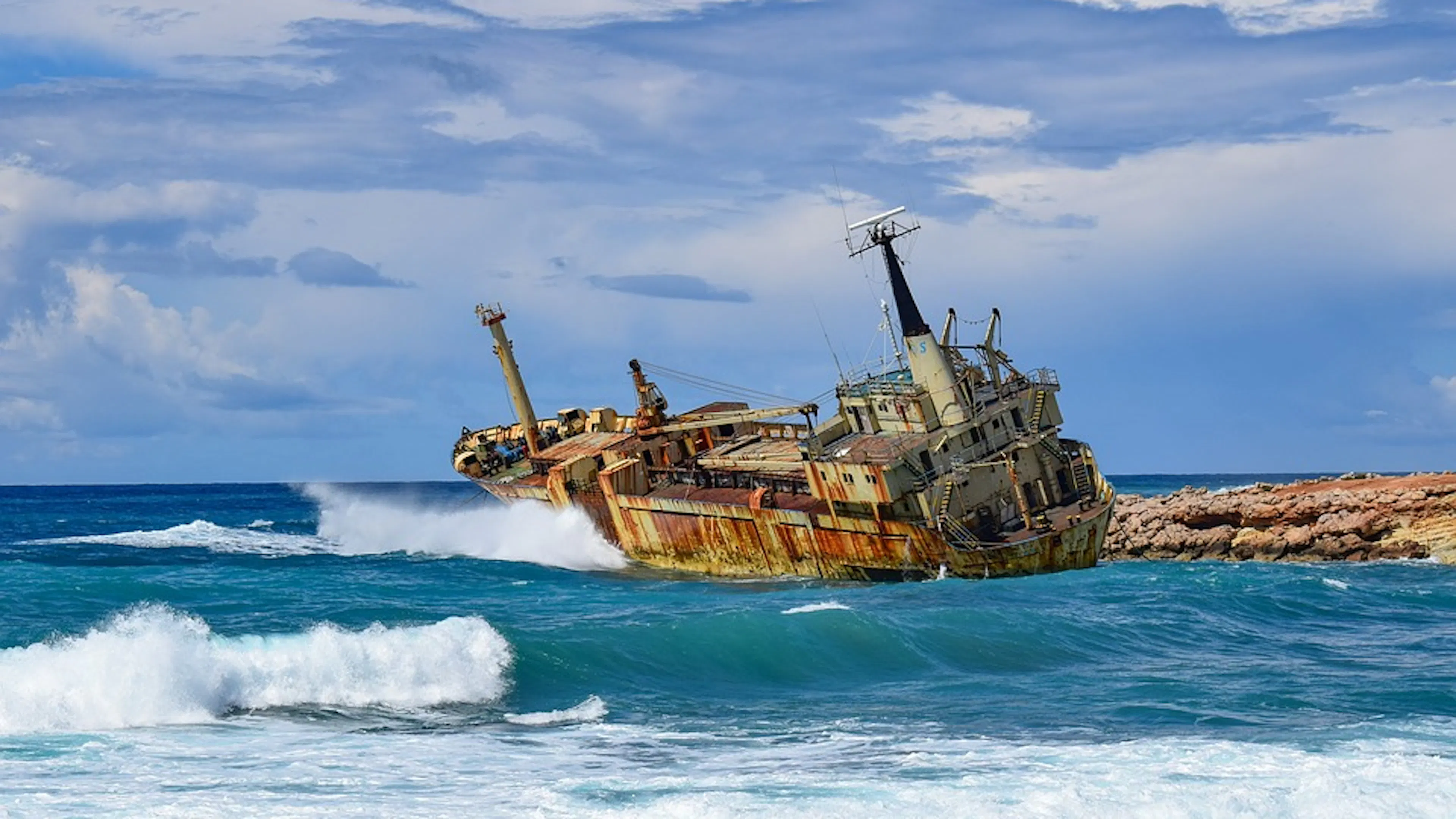 The rusted hull of a shipwreck lodged on some ocean rocks with crashing waves all around it
