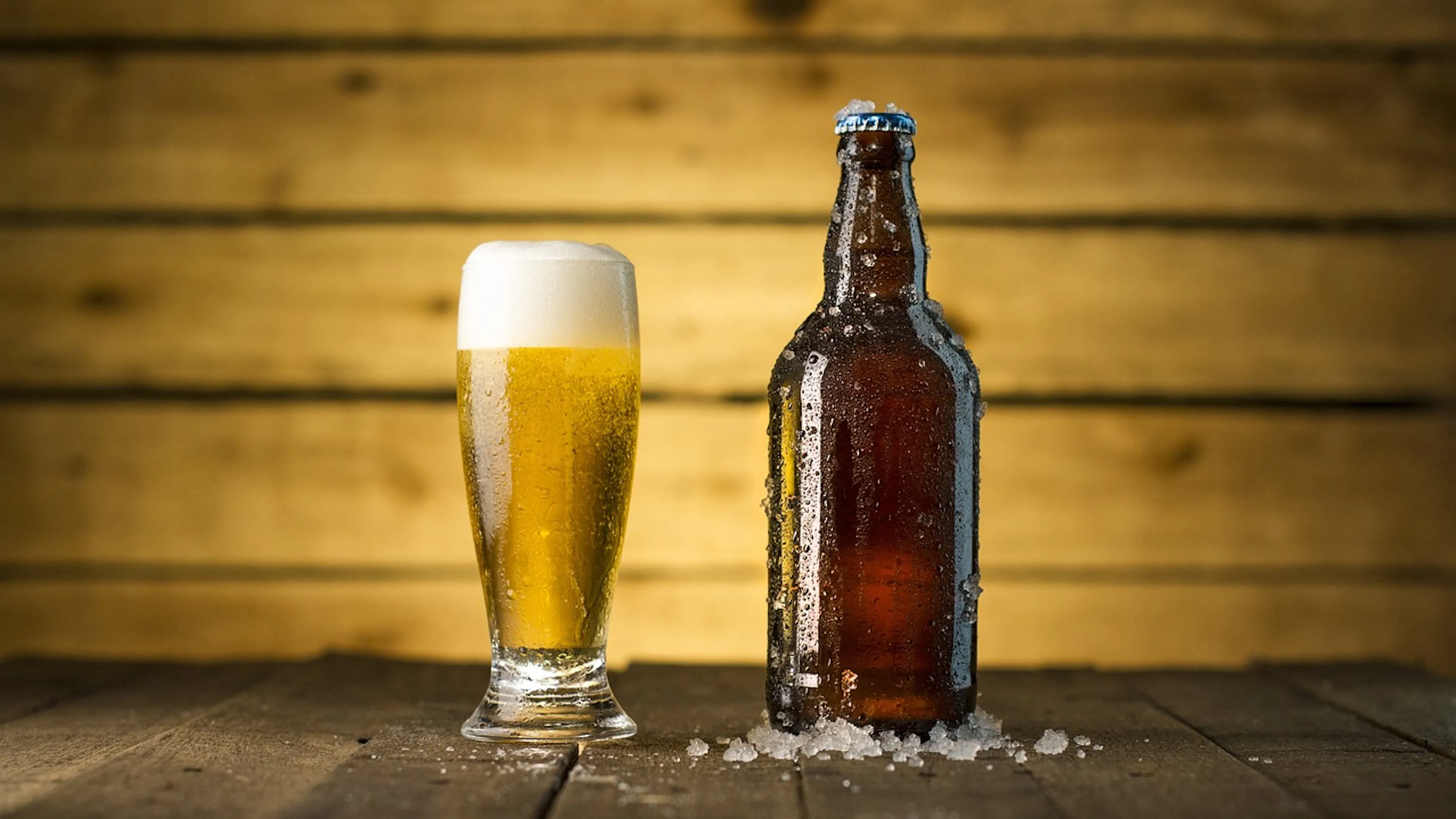 A beer glass and a brown beer bottle side by side in front of a wooden background both filled with a golden beer