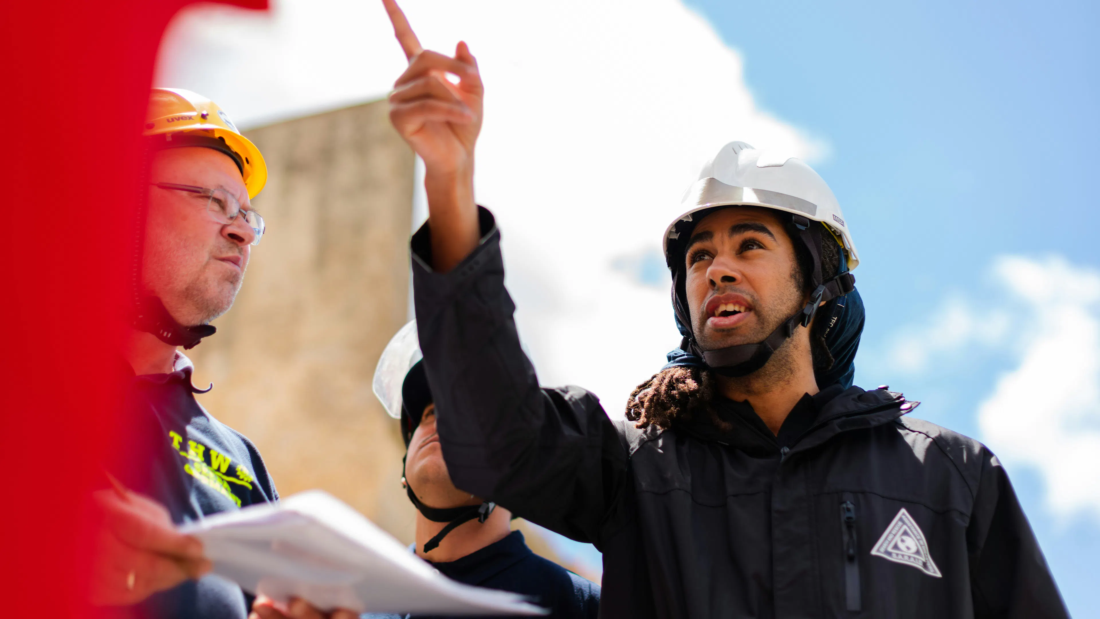 A person wearing a white hard hat pointing toward a construction site with people listening to him talk