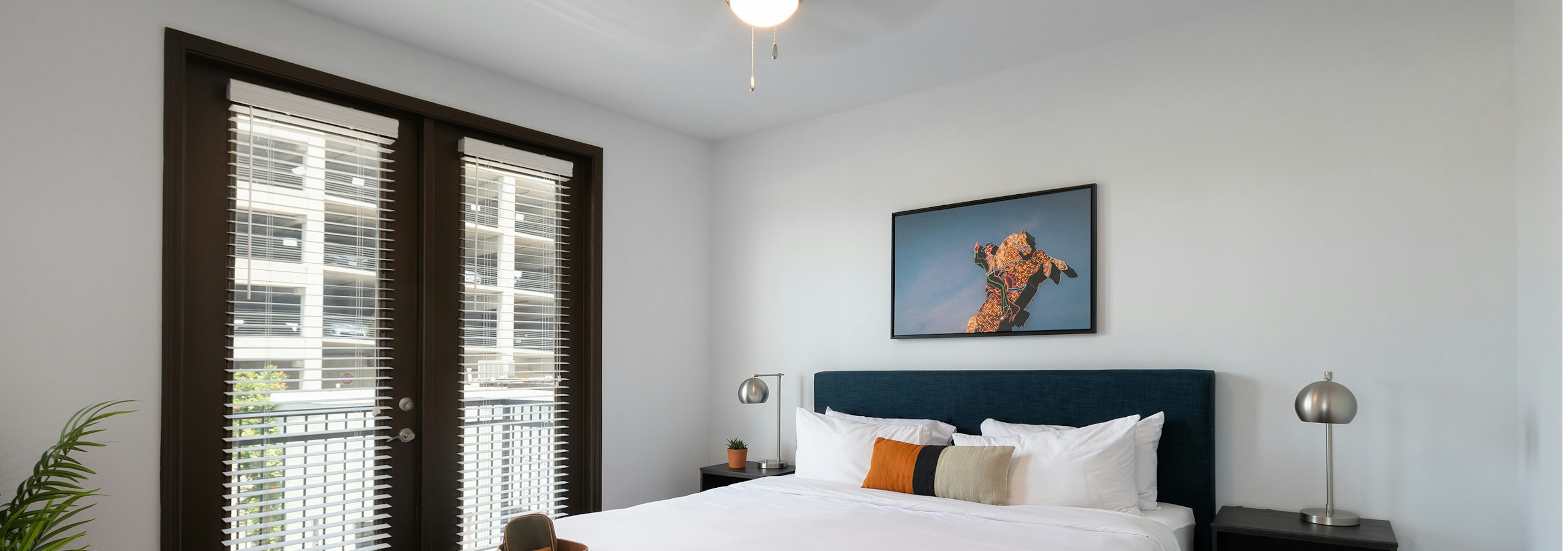 Interior view of AMLI 300 apartment bedroom with private balcony, large windows with shades, and a ceiling fan