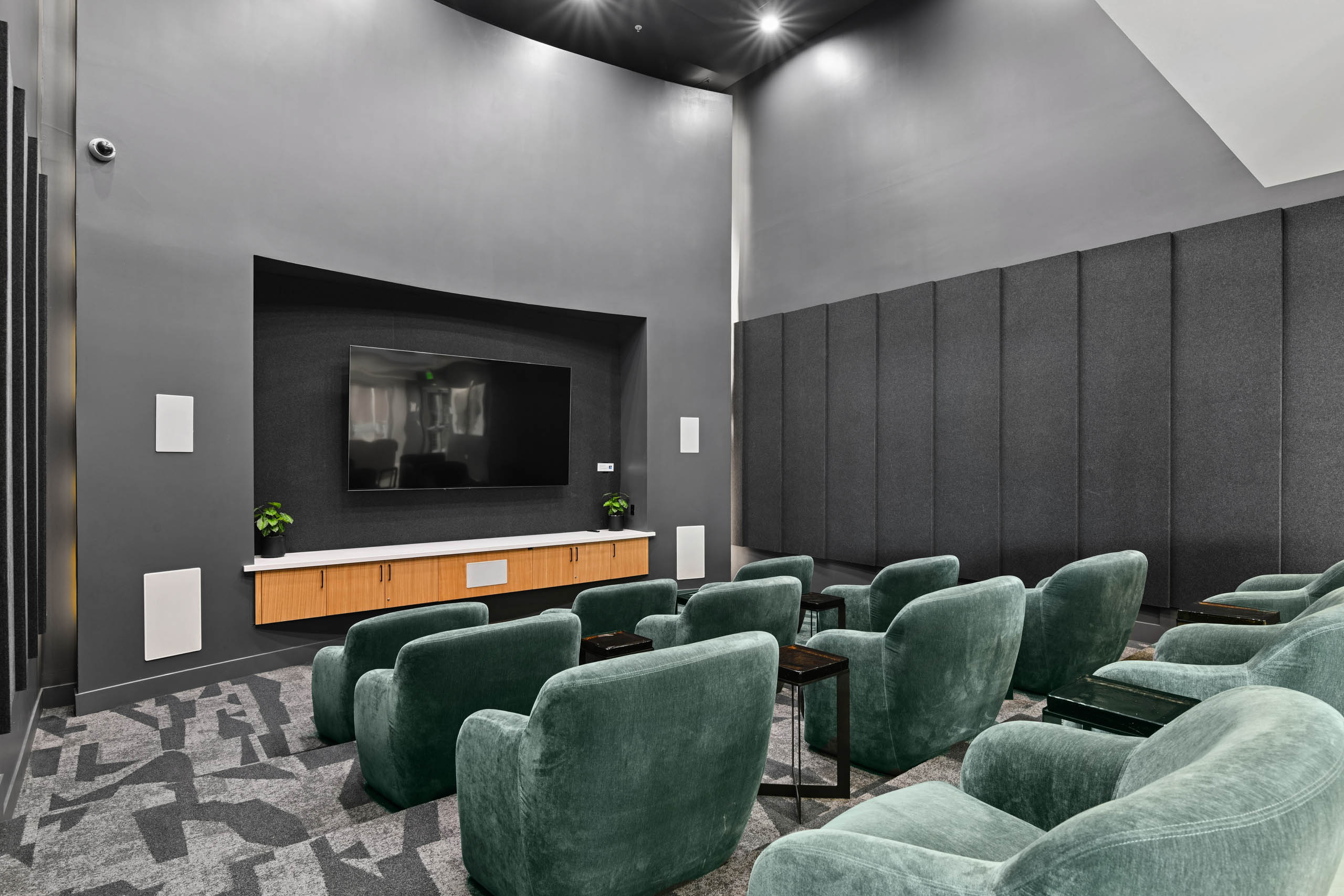 Media lounge at AMLI Lex on Orange with rows of green velvet armchairs, small side tables, a large wall-mounted TV above wood cabinetry, dark gray walls with padded wall panels, and patterned carpet.