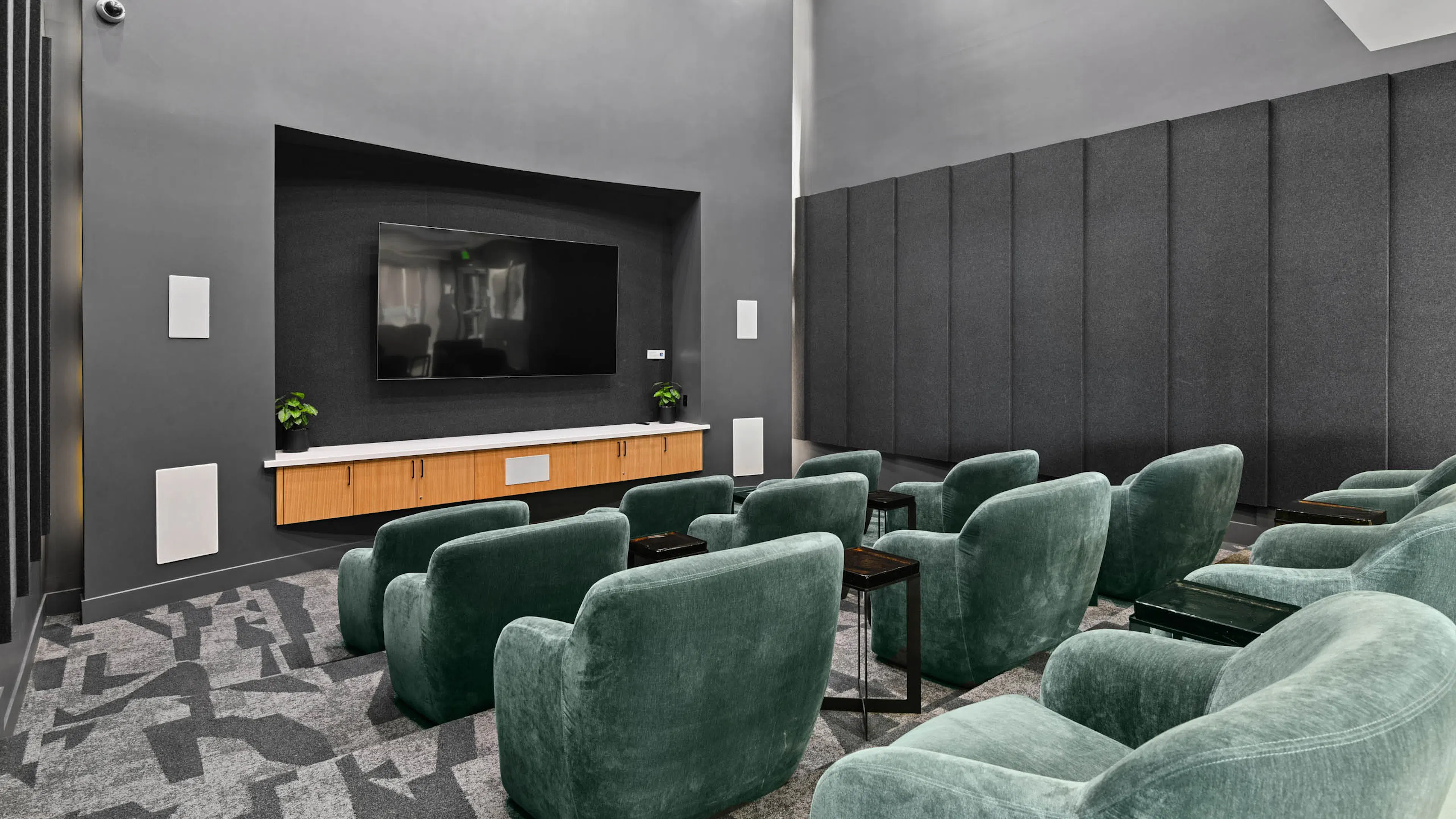 Media lounge at AMLI Lex on Orange with rows of green velvet armchairs, small side tables, a large wall-mounted TV above wood cabinetry, dark gray walls with padded wall panels, and patterned carpet.