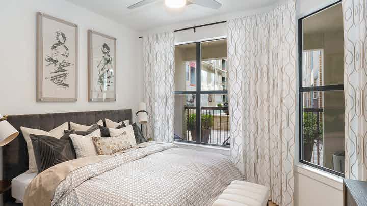 Bedroom at AMLI Quadrangle with a short grey headboard with white bedding and white walls with 2 large windows with blinds