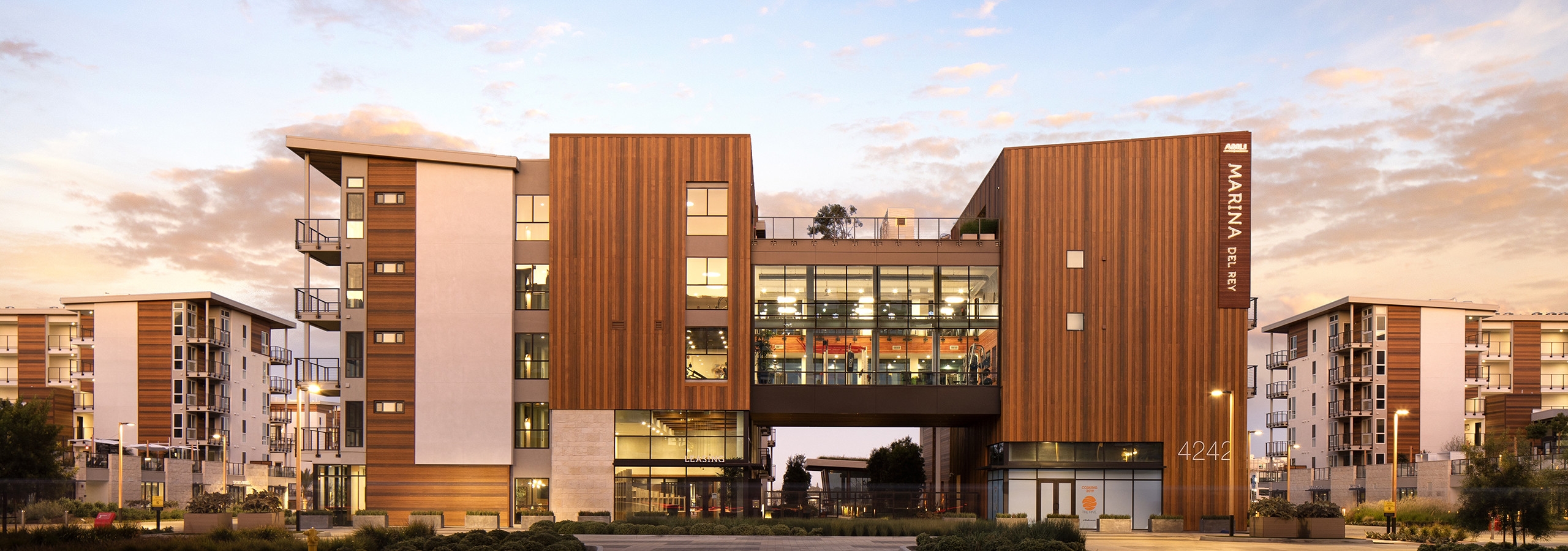 Exterior daytime rendering of AMLI Marina Del Rey apartment building's front entrance wood-paneling facade and lush landscape