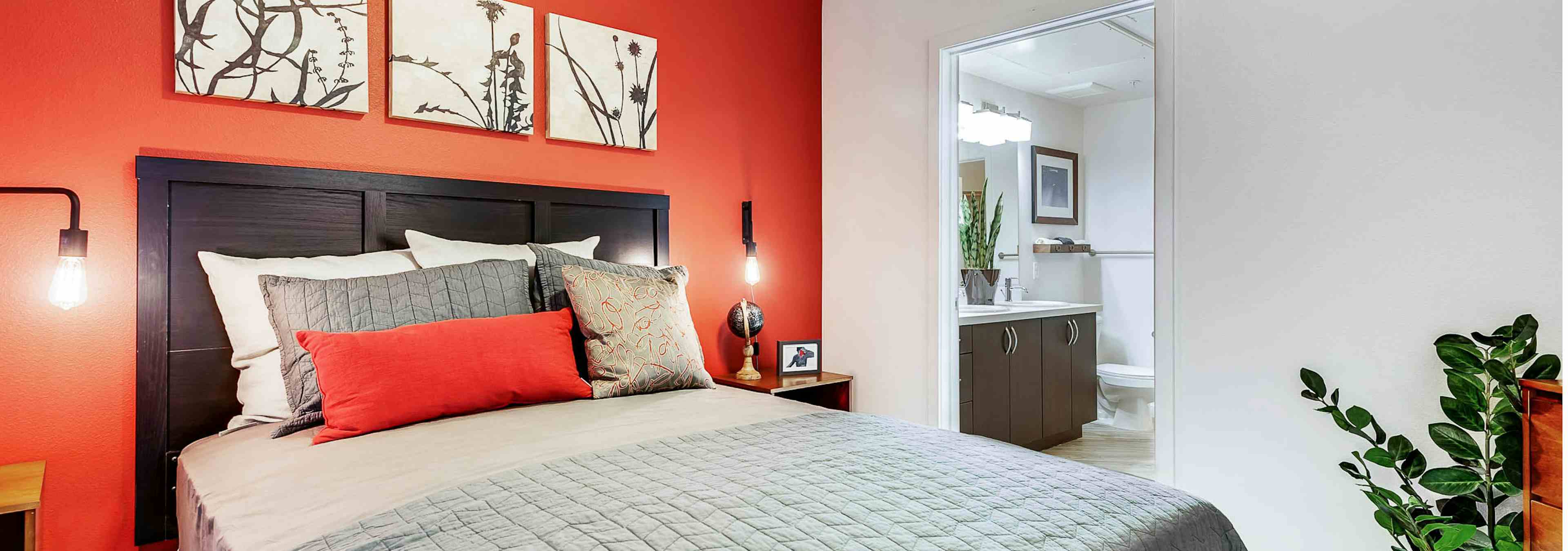 Interior view of a bedroom at AMLI Cherry Creek apartments with a bed backing up to a bright orange wall and view of bathroom