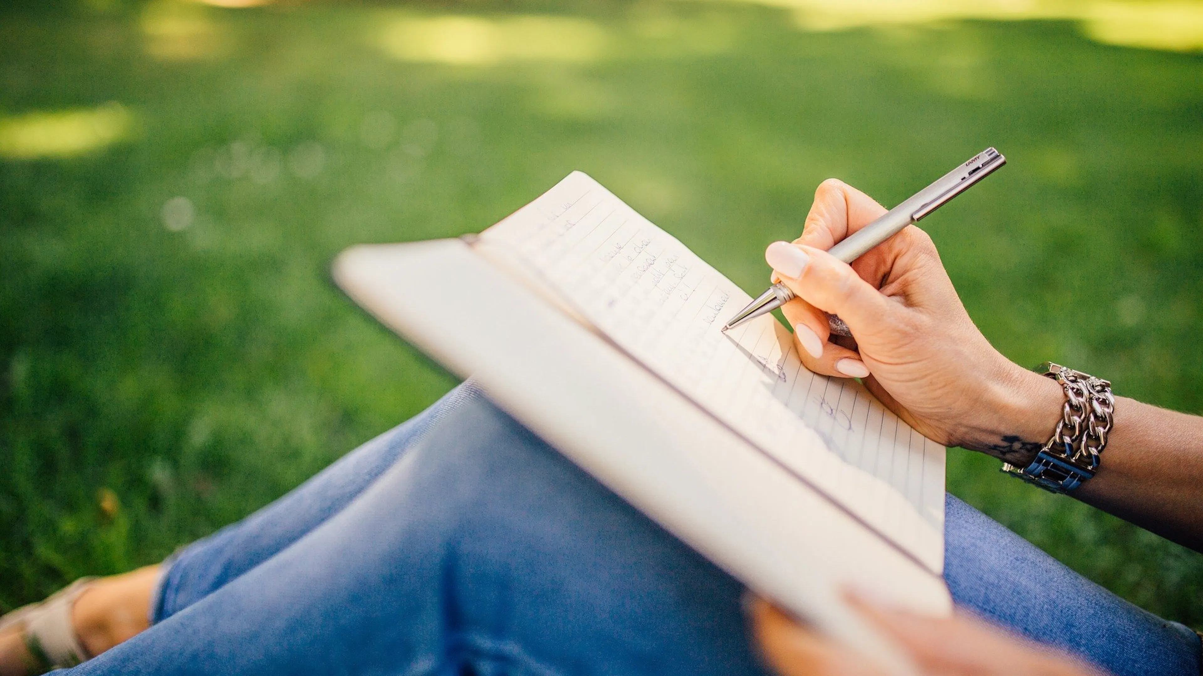 Hand of woman writing in notebook on lap sitting in green grass