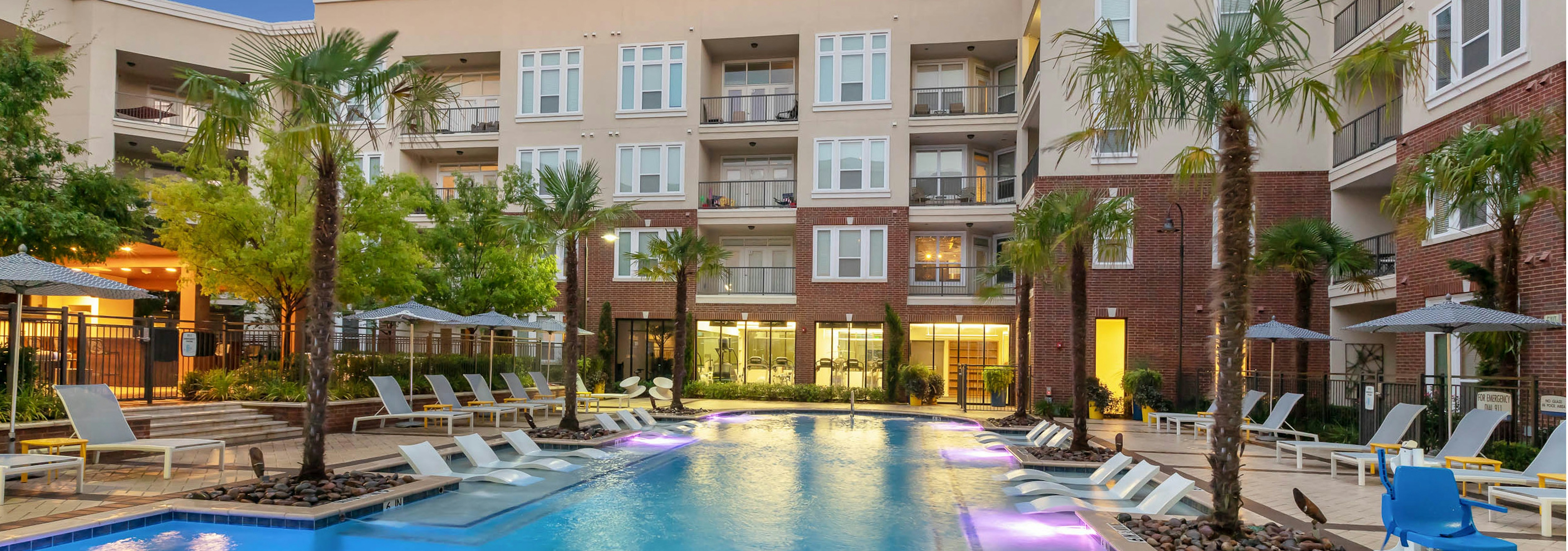 Dusk exterior view of pool area at AMLI Frisco Crossing apartments with lounge seating and umbrellas and palm trees