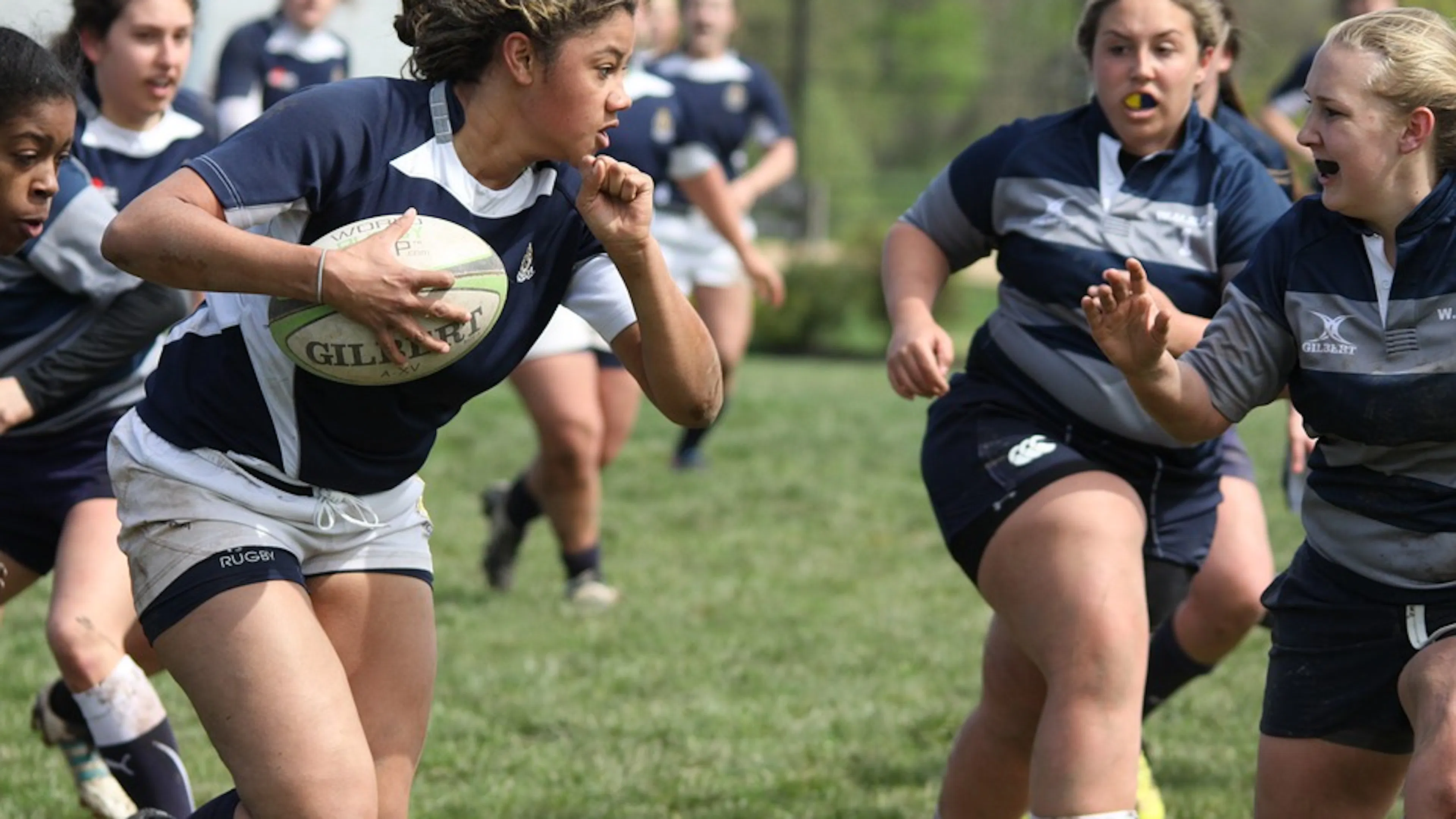 Women playing rugby in black and white striped uniforms running with a rugby ball on a grass field