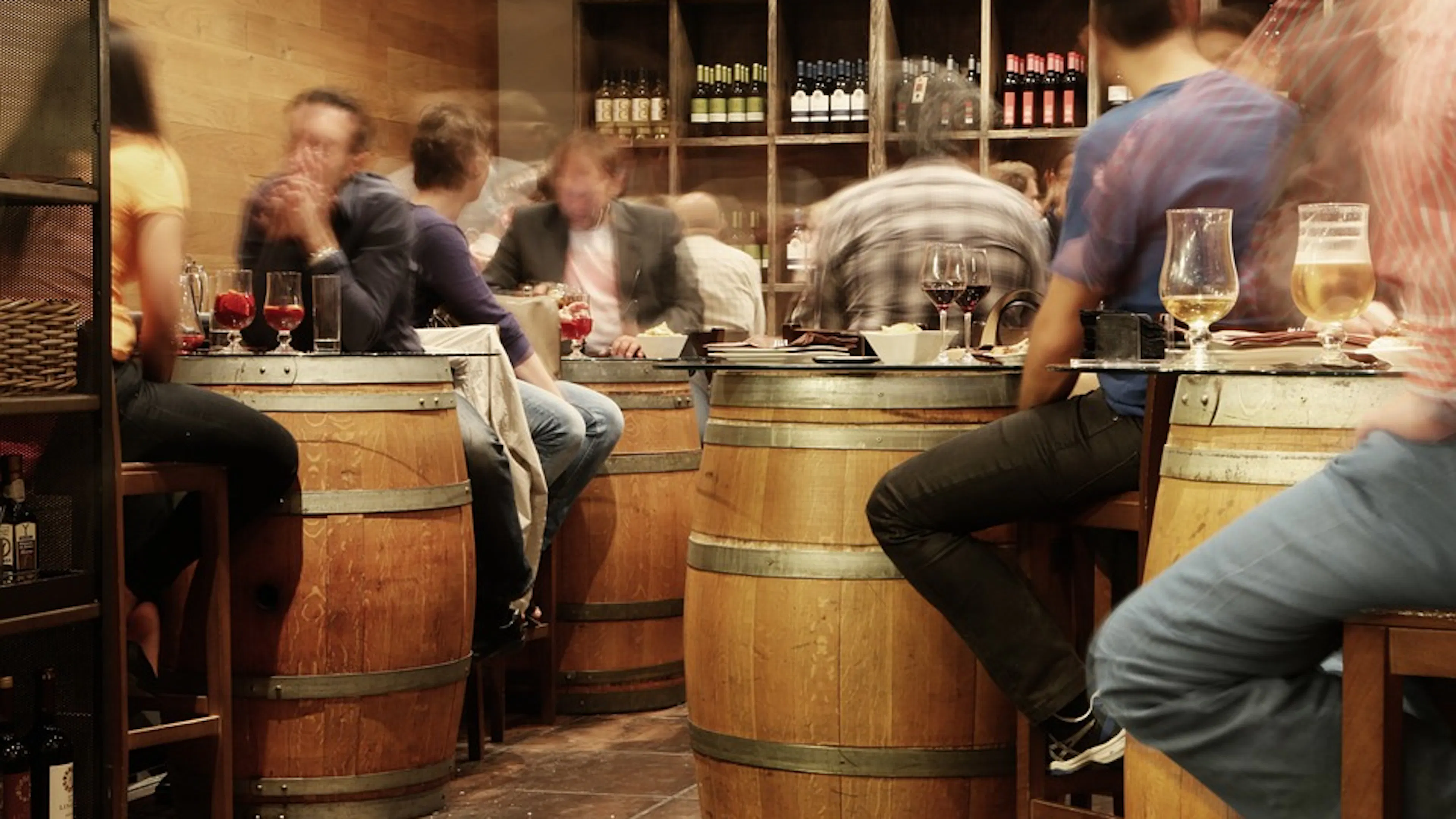 An indoor brewery with upright barrels for tables and a time lapse streak of people moving around