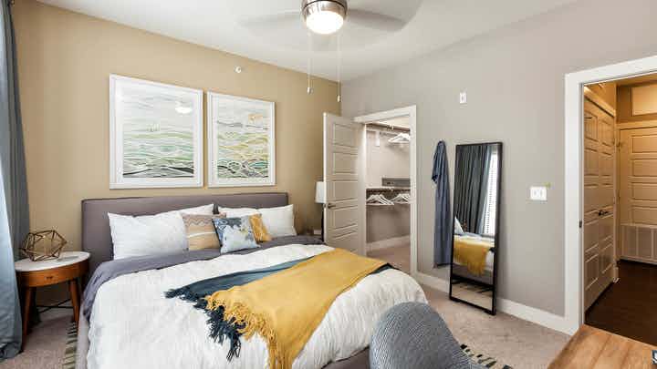 Interior of AMLI West Plano bedroom with tan walls and a white bed with a yellow throw blanket and a peek into walk in closet