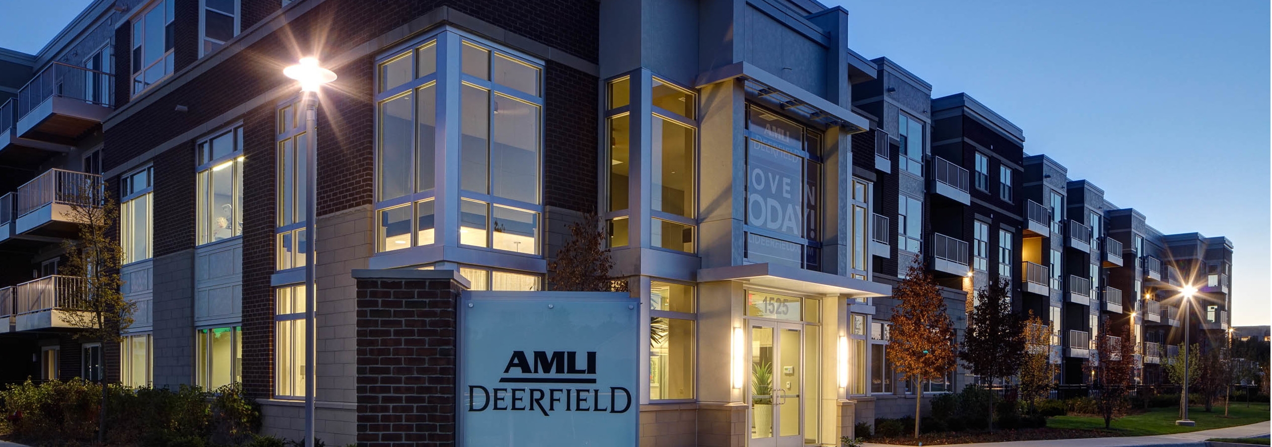 Dusk exterior view of the corner of the AMLI Deerfield main building with landscaping and interior lights beaming