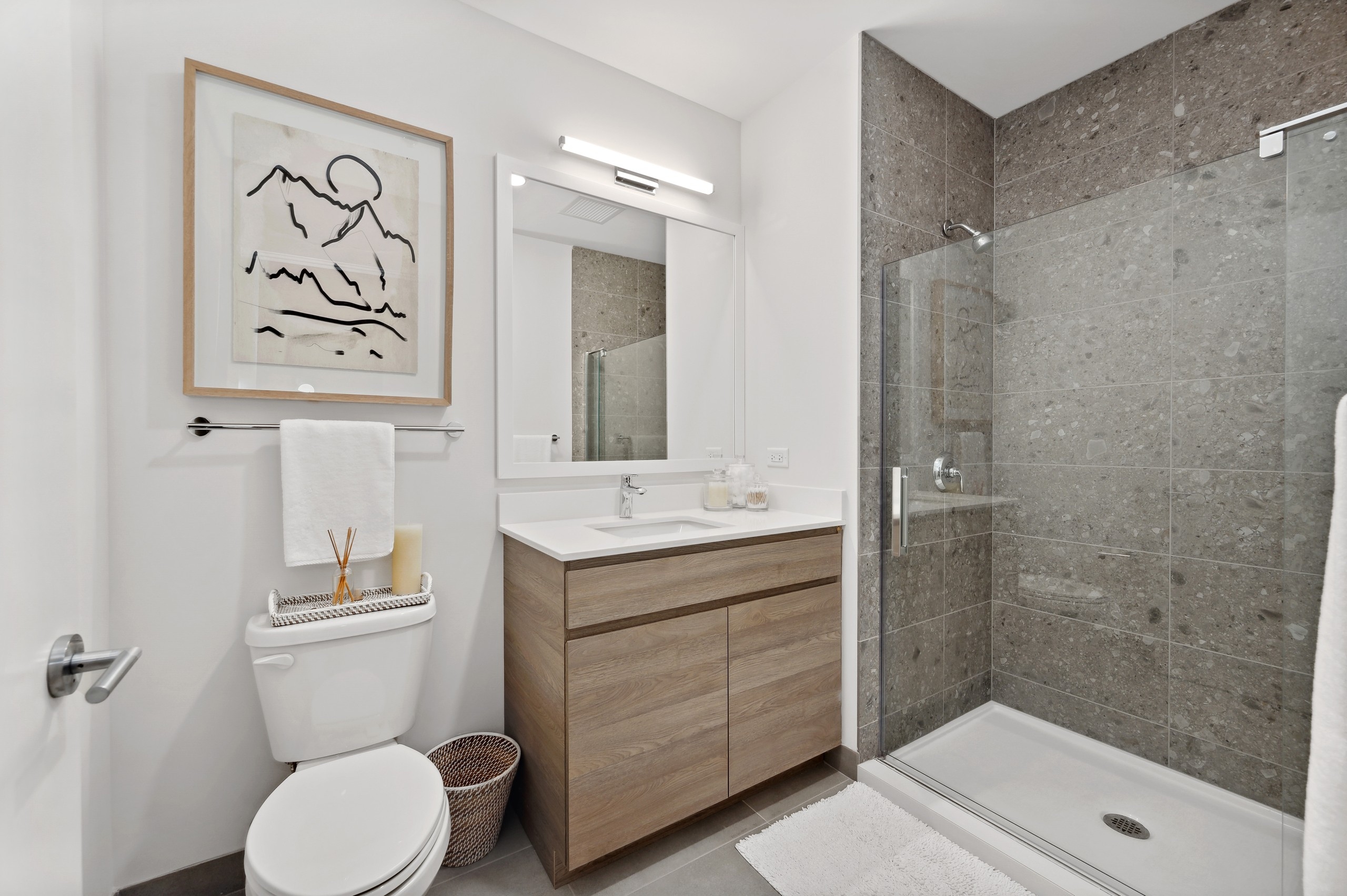 Bathroom at AMLI 808 with black and white artwork and light wood cabinet and full glass enclosed shower with oversized tile