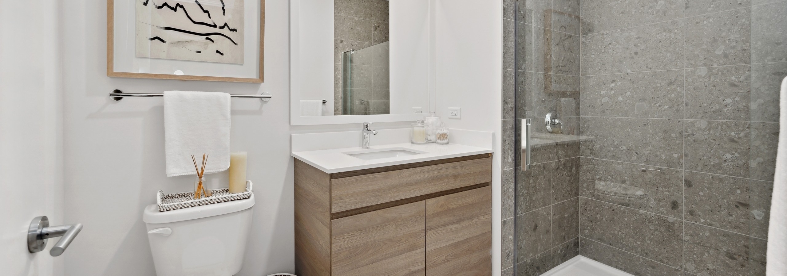 Bathroom at AMLI 808 with black and white artwork and light wood cabinet and full glass enclosed shower with oversized tile