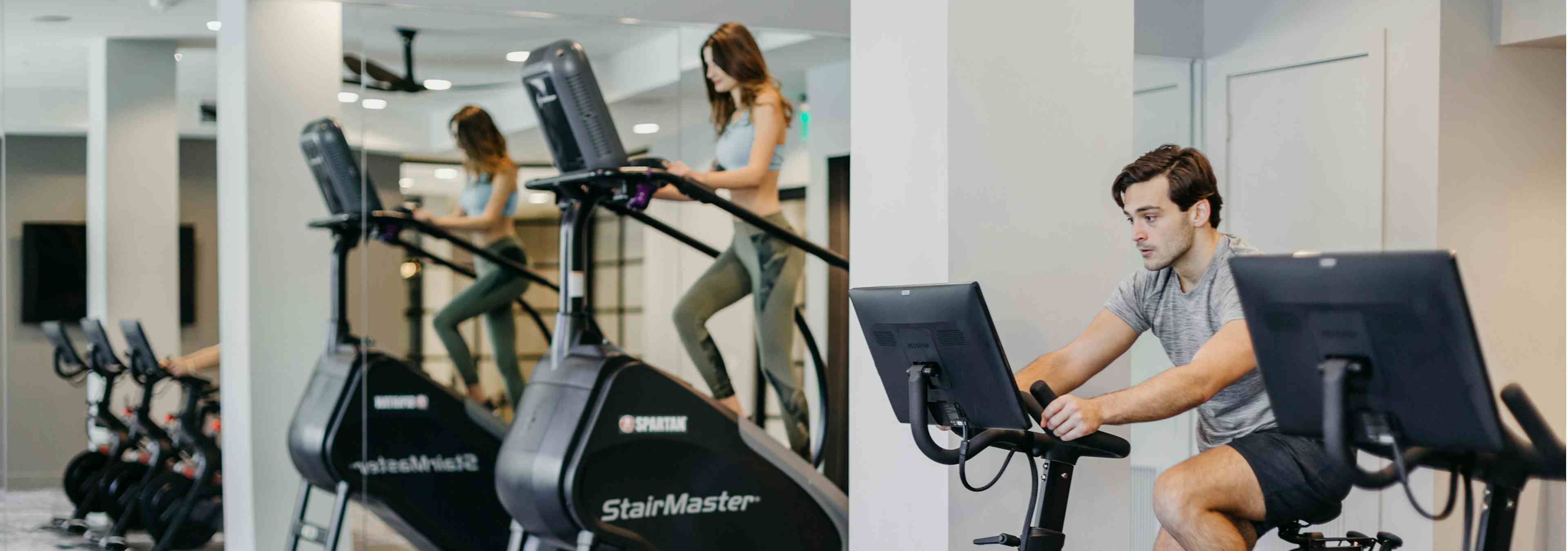 Close up of AMLI Addison resident using a Peloton bike and two residents using stair climbing machines in the fitness center