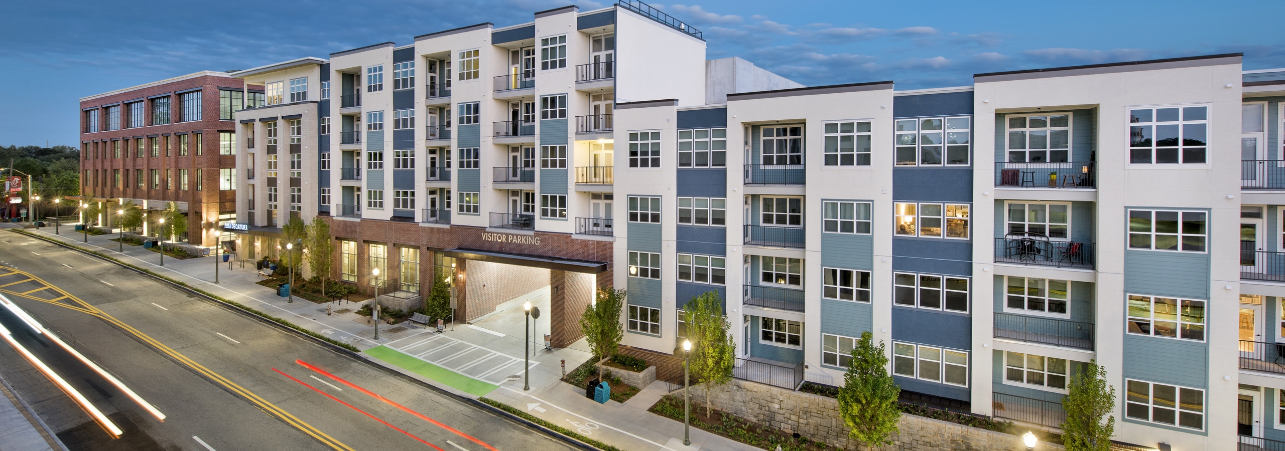 Dusk view of the side of the AMLI Decatur apartment building with the parking entrance and lamp posts and manicured trees