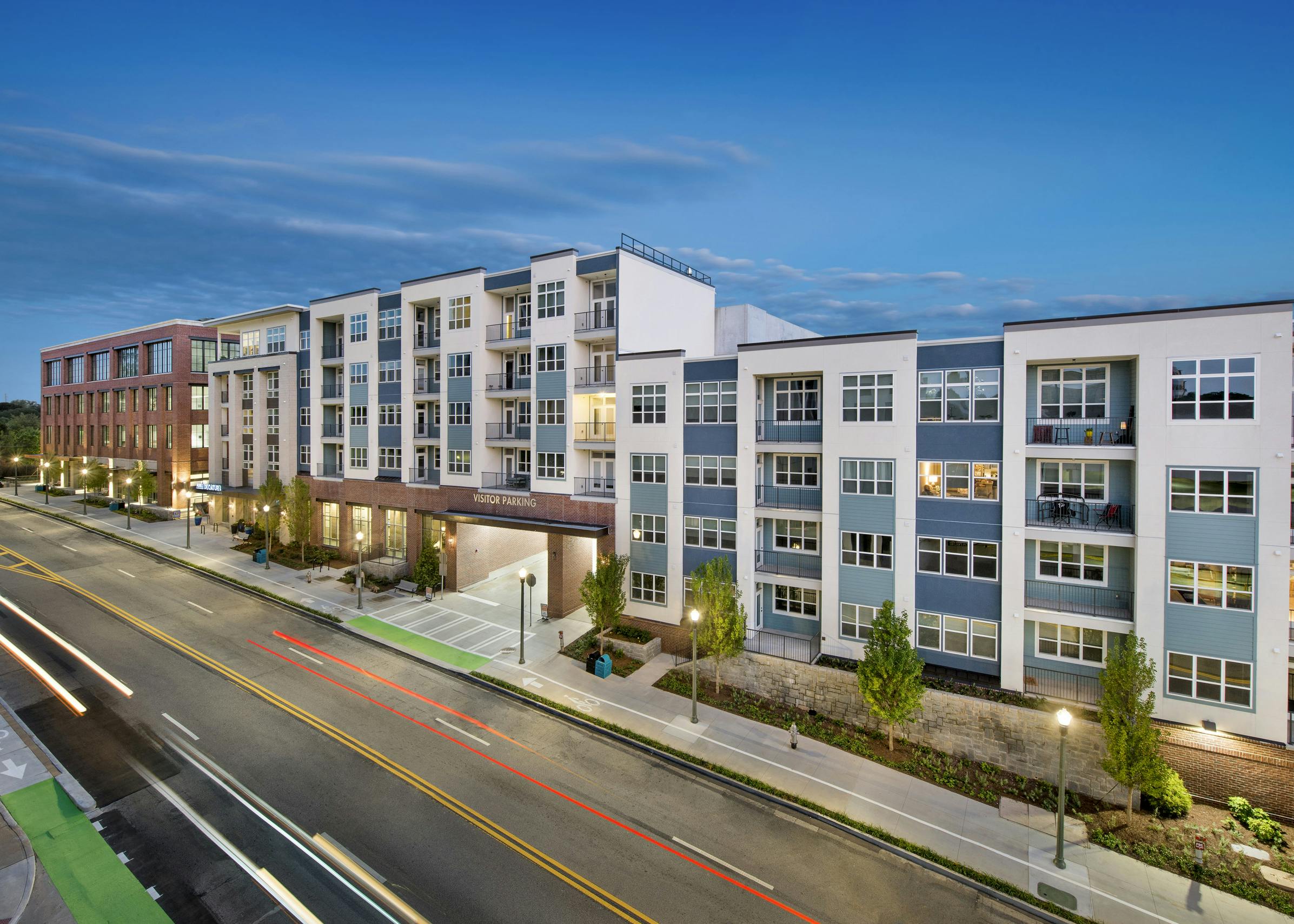 Dusk view of the side of the AMLI Decatur apartment building with the parking entrance and lamp posts and manicured trees