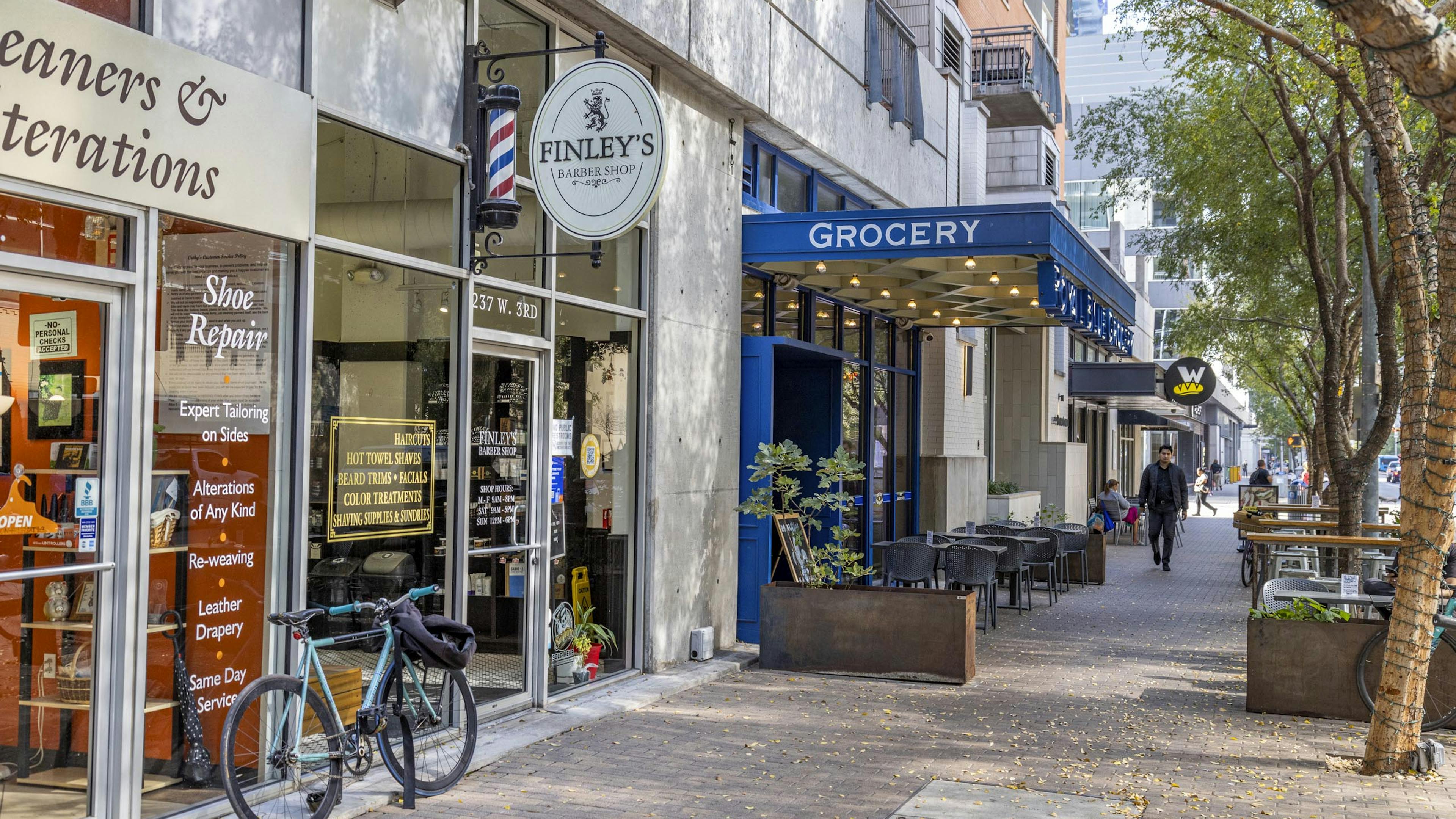 Daytime view of AMLI Downtown retailers including a barber shop and a grocery store with blue signage and outdoor seating