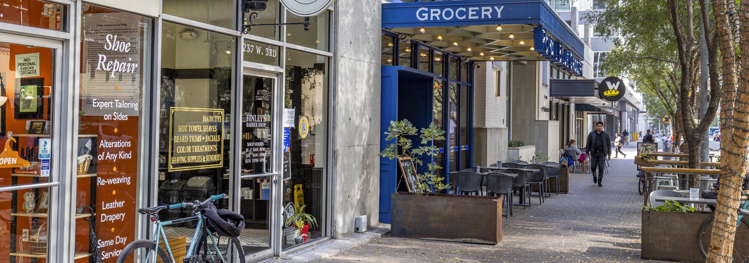 Daytime view of AMLI Downtown retailers including a barber shop and a grocery store with blue signage and outdoor seating