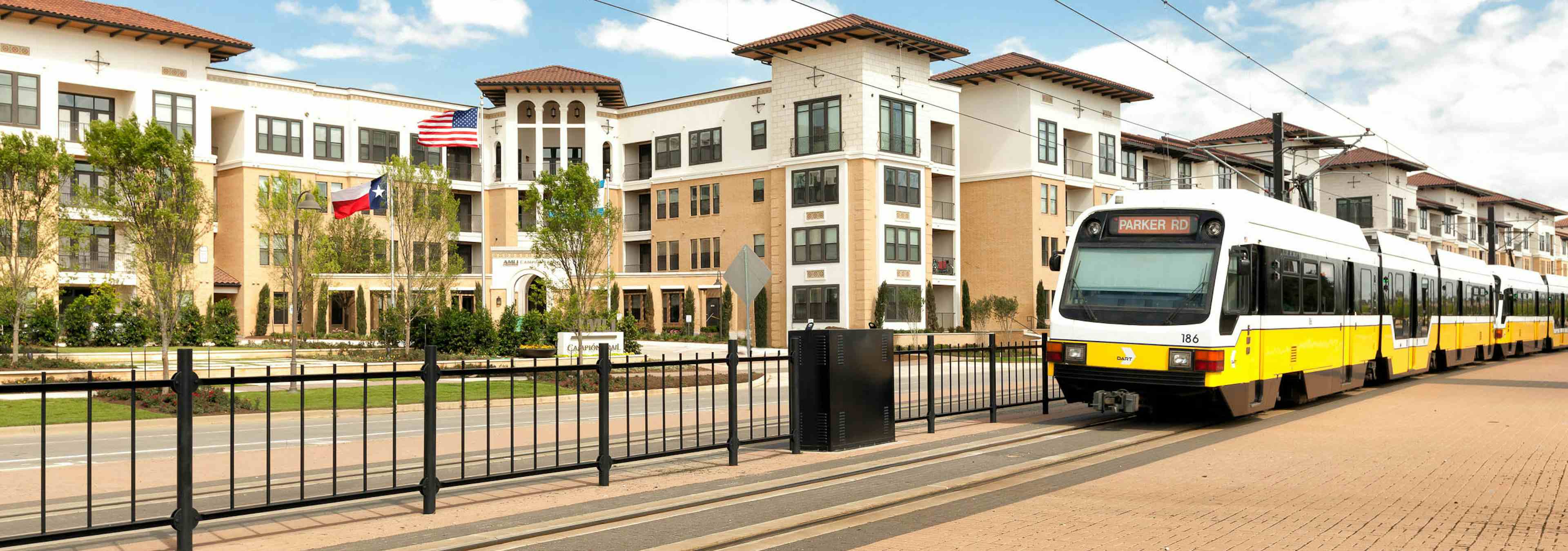 Daytime view of light rail train on tracks along fence traveling in front of the AMLI Campion Trail apartment building