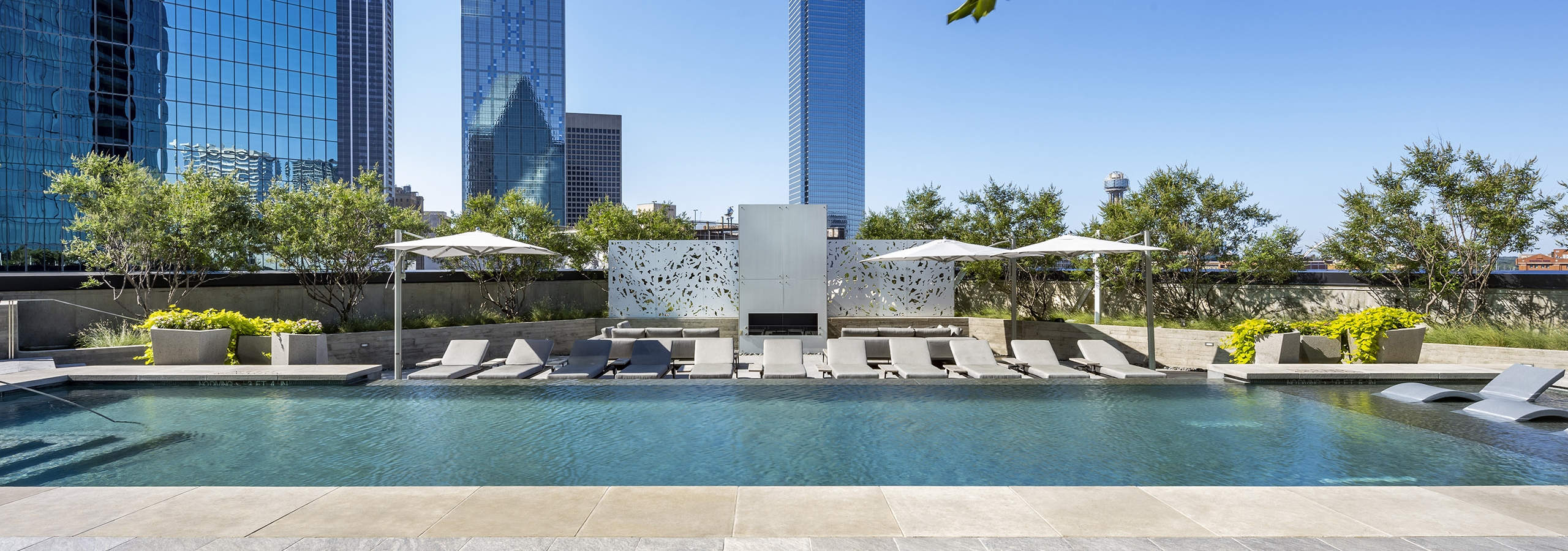 AMLI Fountain Place pool surrounded by grey stones deck with grey lounge chairs and fireplace in front of city skyline