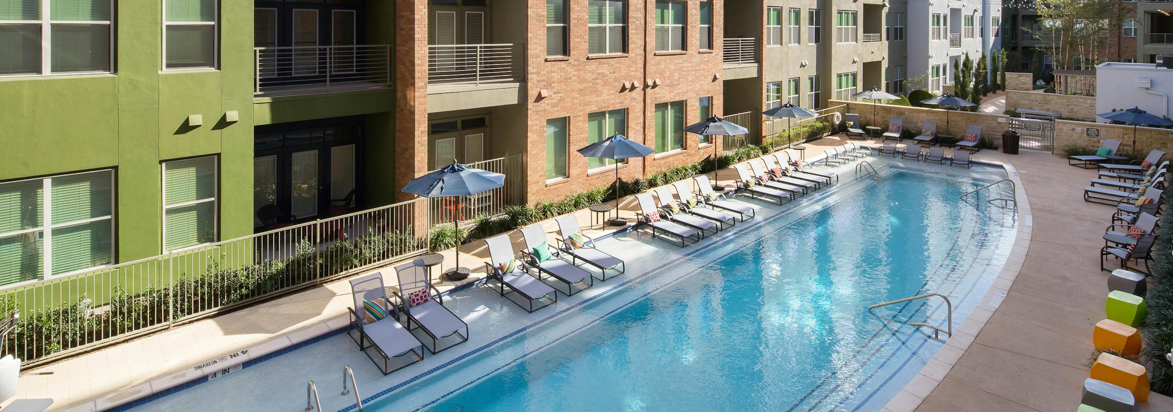 Exterior view of pool at AMLI at Mueller with blue umbrellas and white lounge chairs with facing building facade