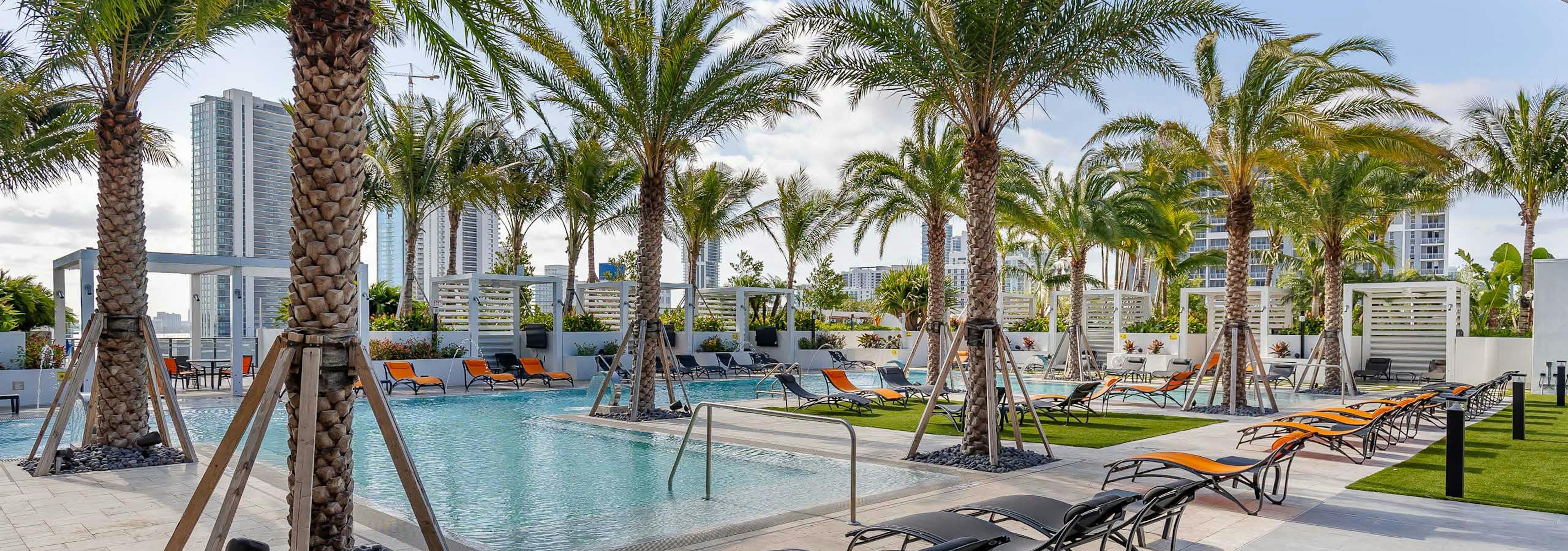 Daytime view of the AMLI Midtown Miami pool deck with orange and black lounge chairs and palm trees and a view of the city 