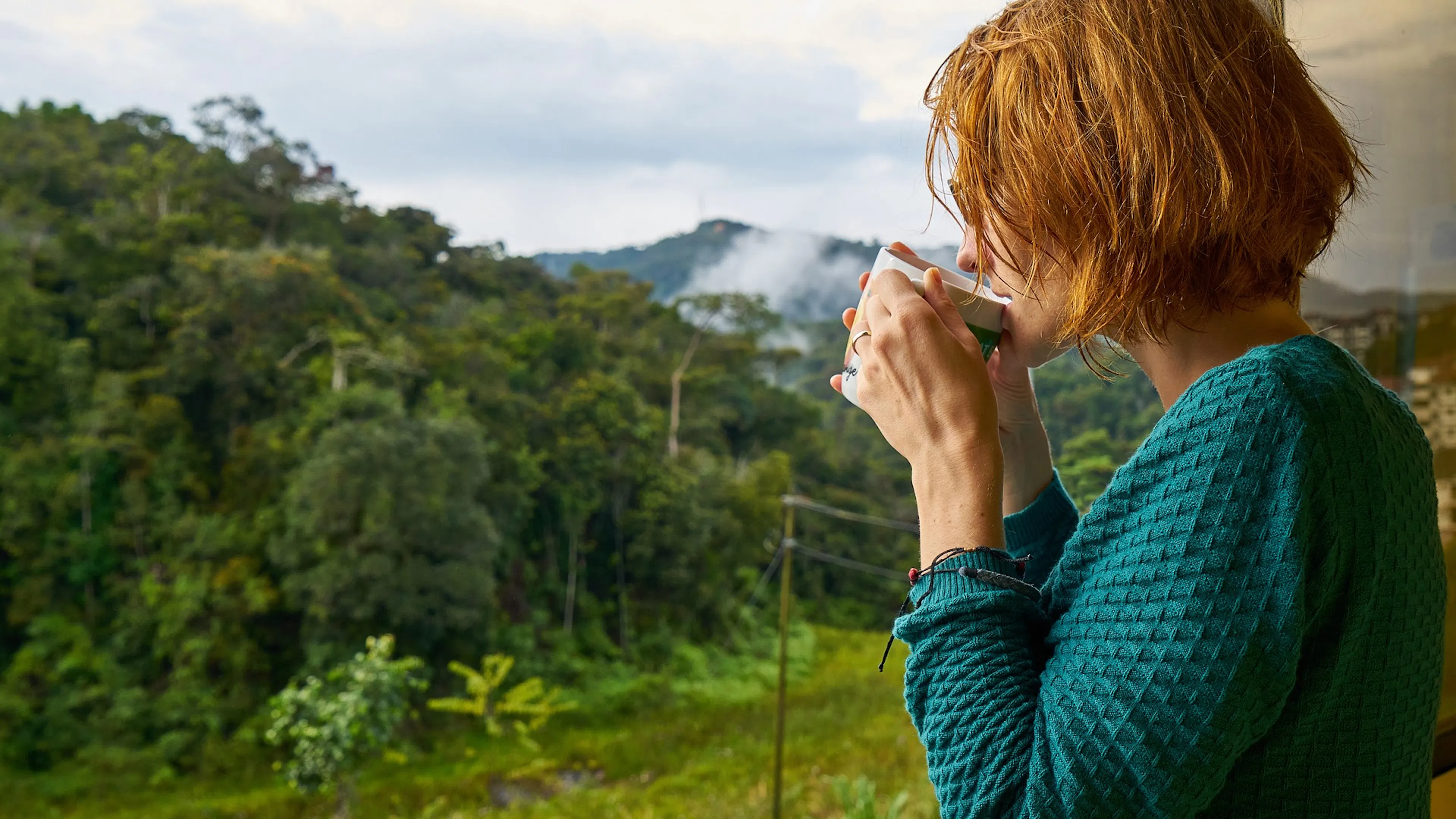 Woman with short red hair in blue sweater holding a cup of coffee looking out at green trees