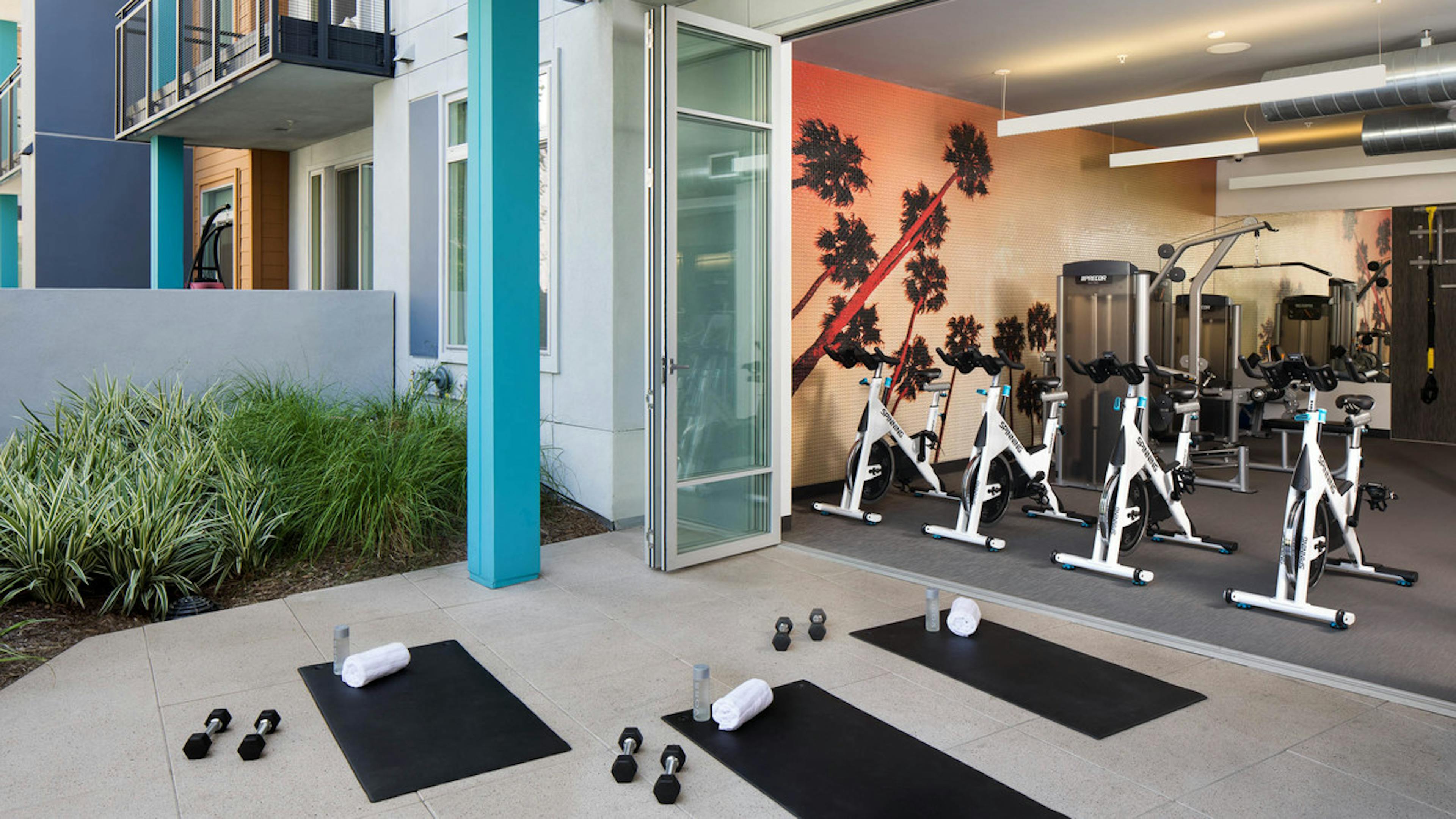 Fitness center at AMLI Uptown Orange apartments with spin bikes overlooking outdoor exercise deck, yoga mats and dumbbells